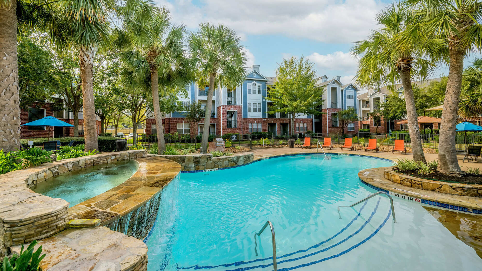 Resort-style swimming pool with a stone waterfall spa, surrounded by palm trees, orange lounge chairs, and brick apartment buildings under a cloudy sky.