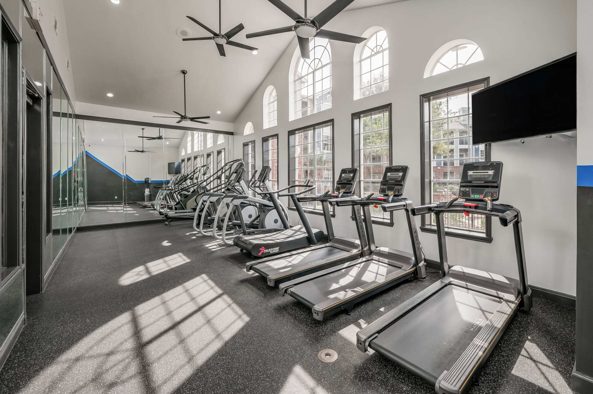 Bright fitness center with vaulted ceilings, ceiling fans, and arched windows, featuring a row of treadmills and elliptical machines on dark flooring with a wall-mounted TV.