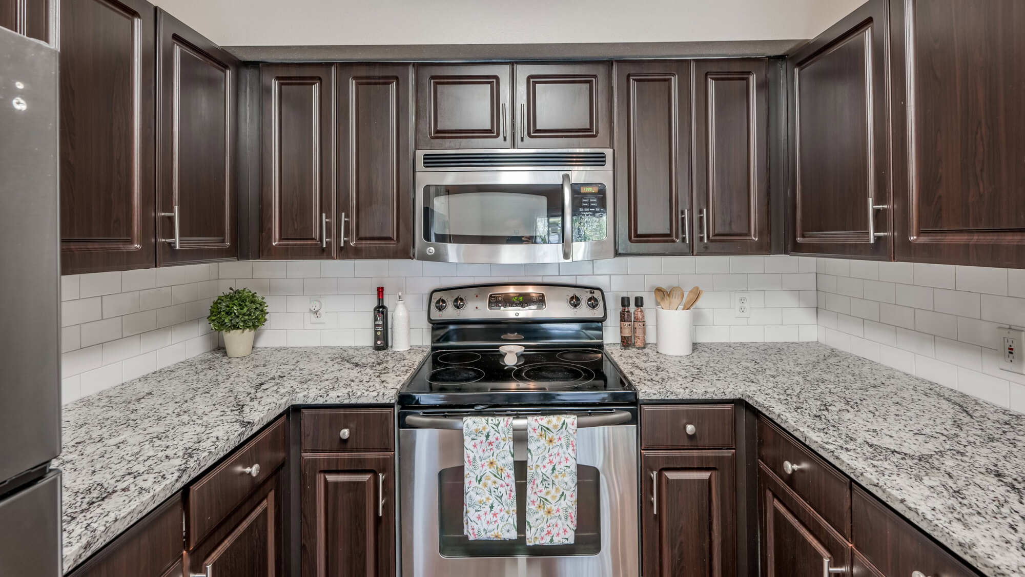 Apartment kitchen with dark wood cabinets, granite countertops, white subway tile backsplash, and stainless steel appliances including a microwave and electric range.