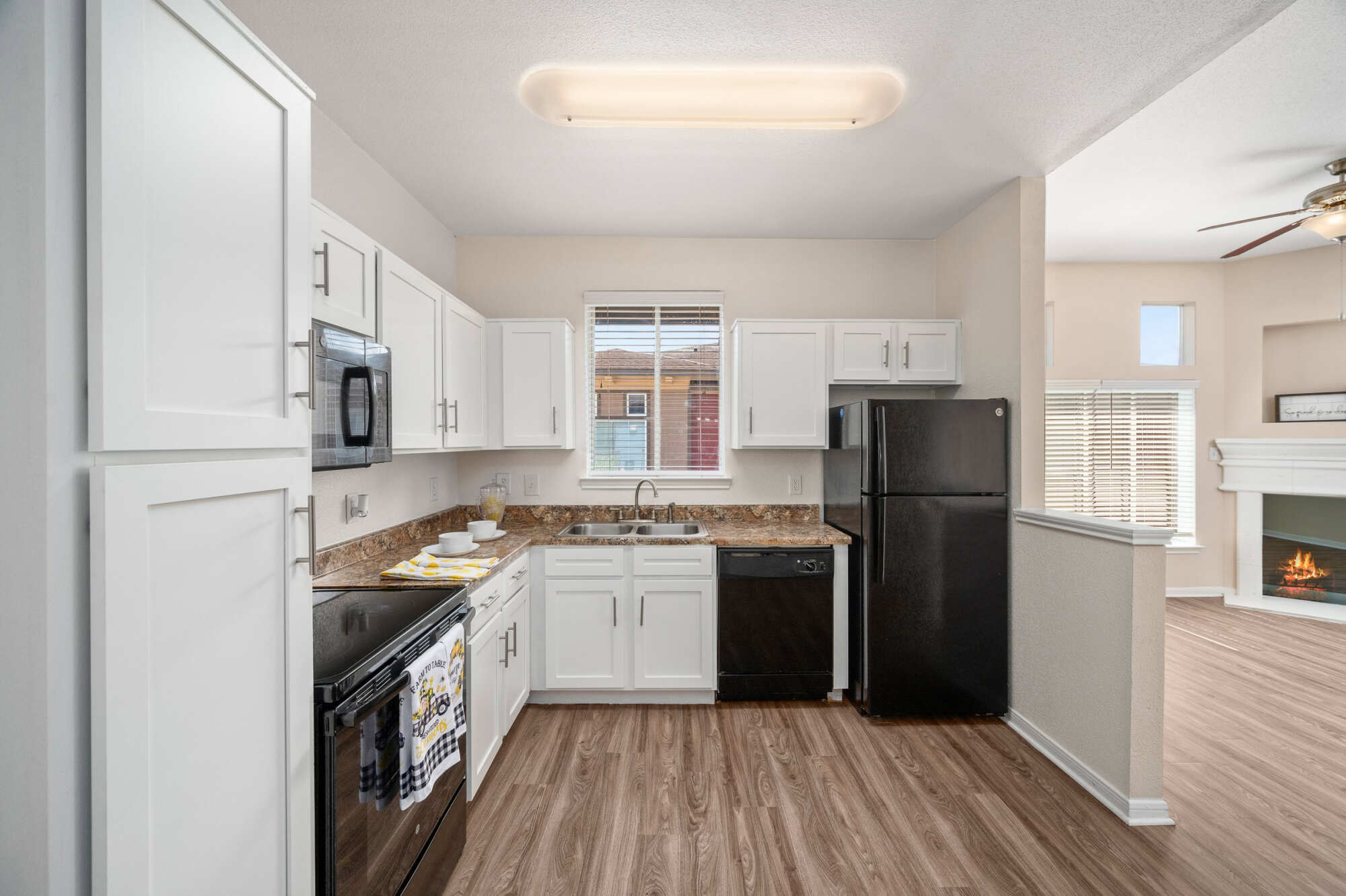 Open apartment kitchen with white cabinetry, granite countertops, black appliances, and a window over the sink, adjacent to a living area with a lit fireplace.