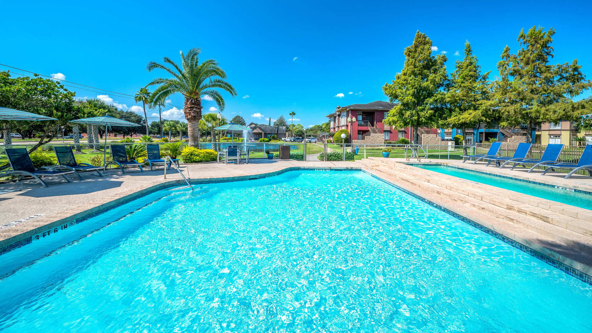 Resort-style swimming pool with crystal-blue water, lounge chairs, shade umbrellas, and palm trees in front of apartment buildings on a sunny day.