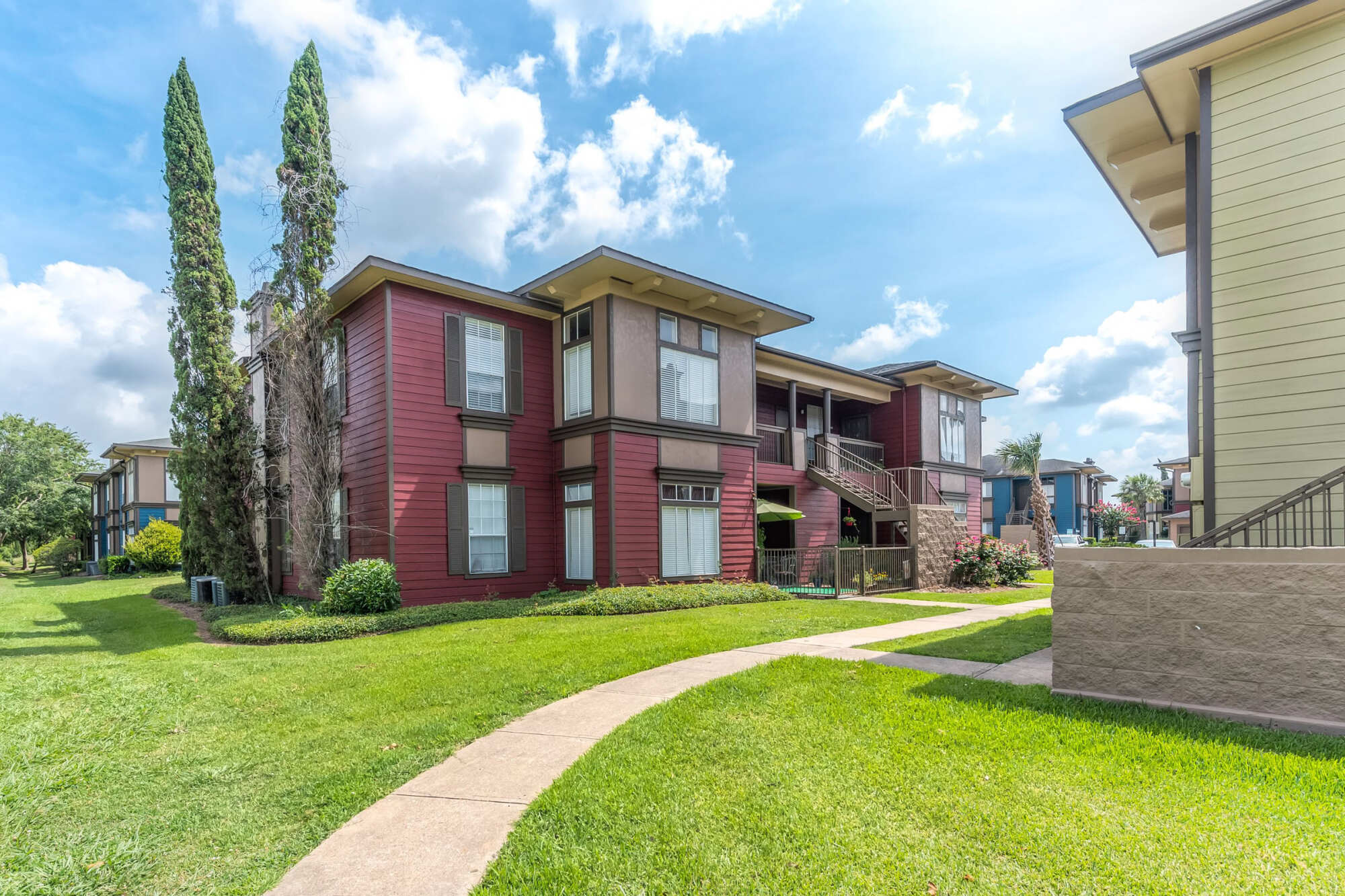 Exterior of a two-story apartment building with red and tan siding, shuttered windows, an exterior staircase, and a curving walkway through a green lawn.