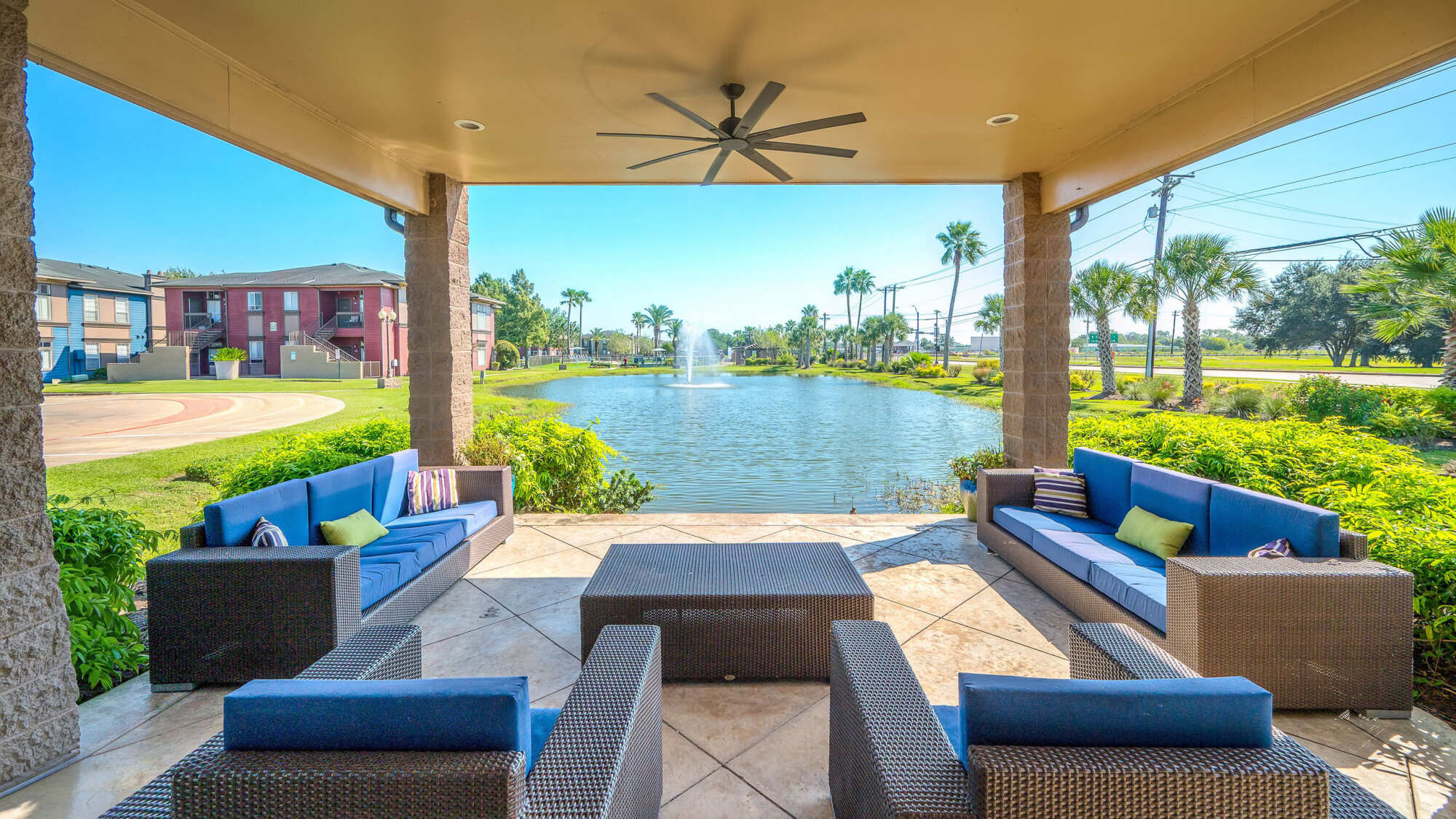 Covered outdoor lounge with wicker sofas and blue cushions overlooking a landscaped pond with a fountain and apartment buildings in the background.