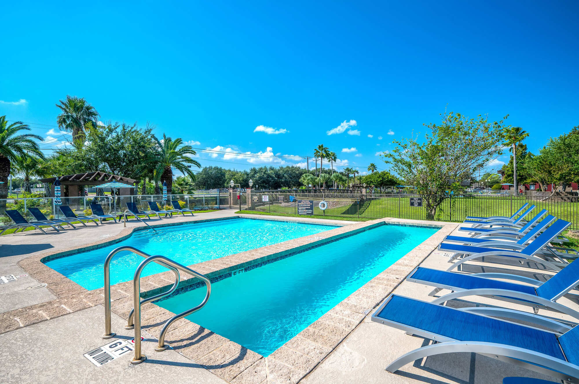 Sparkling blue community pool bordered by a paved deck, blue lounge chairs, palm trees, and a fenced lawn under a clear blue sky.