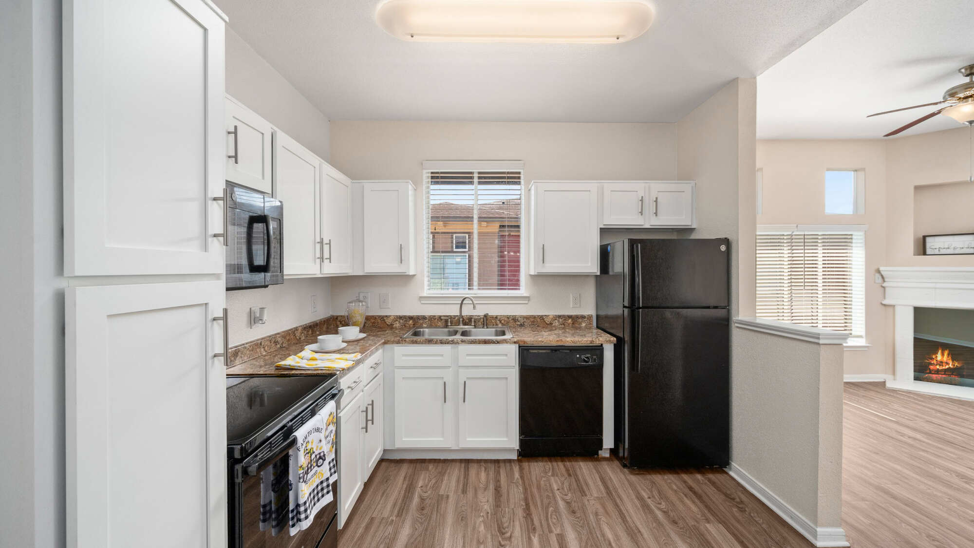 Apartment kitchen with white cabinets, granite countertops, black appliances, and a window over the sink, opening to a living area with a lit fireplace and wood-look flooring.
