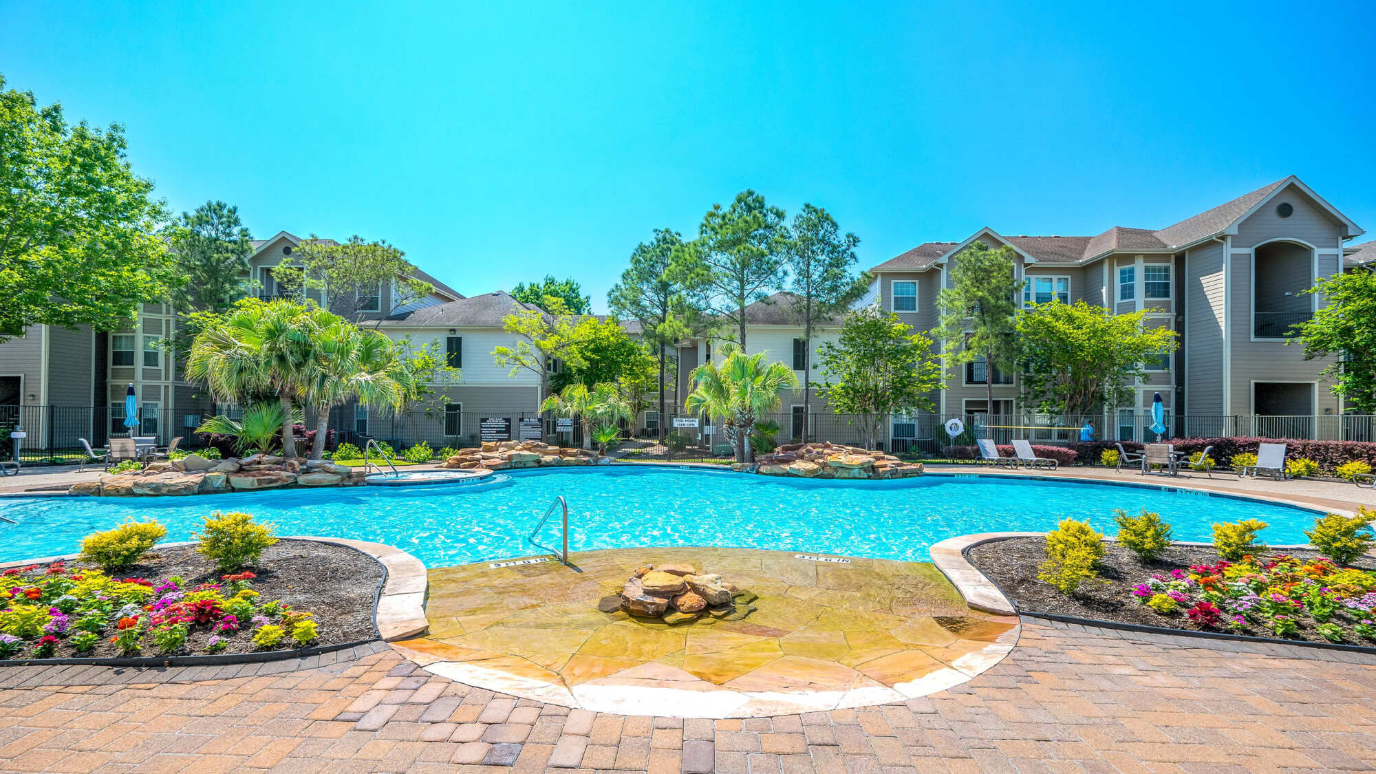 Curving resort-style pool with a mushroom water fountain, clear blue water, palm trees, and tan apartment buildings under a clear sky.