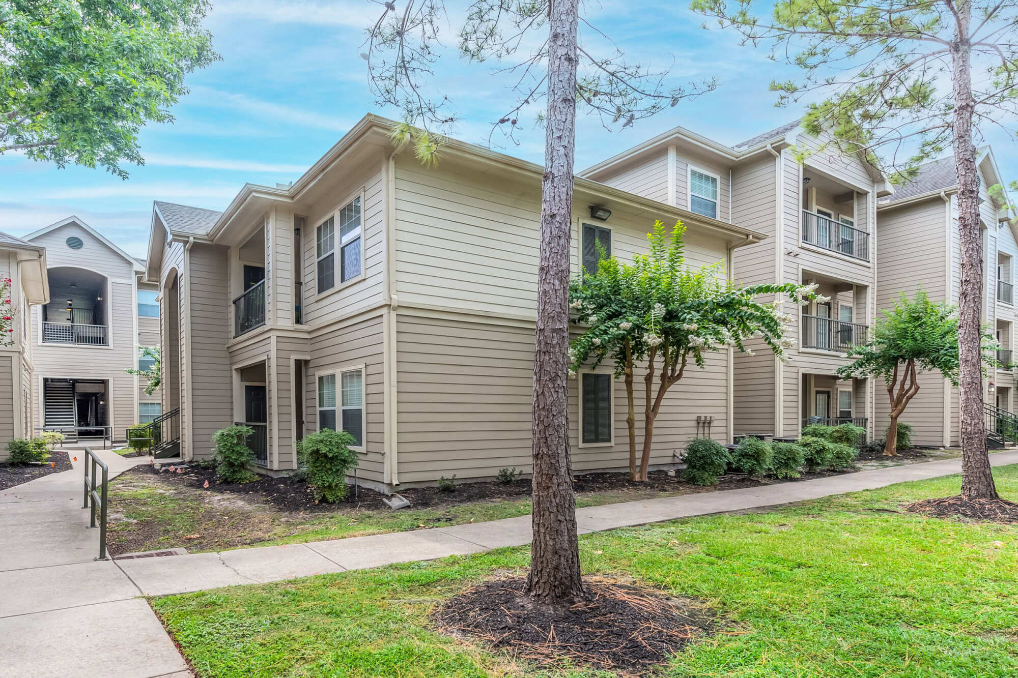 Exterior of a three-story apartment building with tan siding, pitched roofs, and balconies, framed by mature pine trees and landscaped walkways.