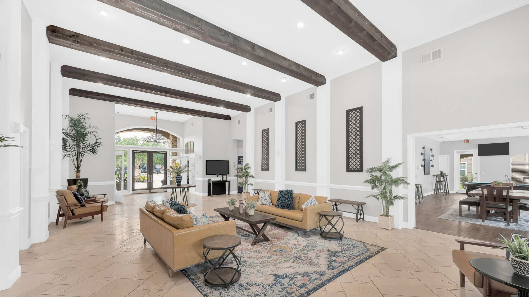 Spacious clubhouse lounge with exposed wood ceiling beams, tan leather sofas, a patterned area rug, tall windows, and a dining area in the background.