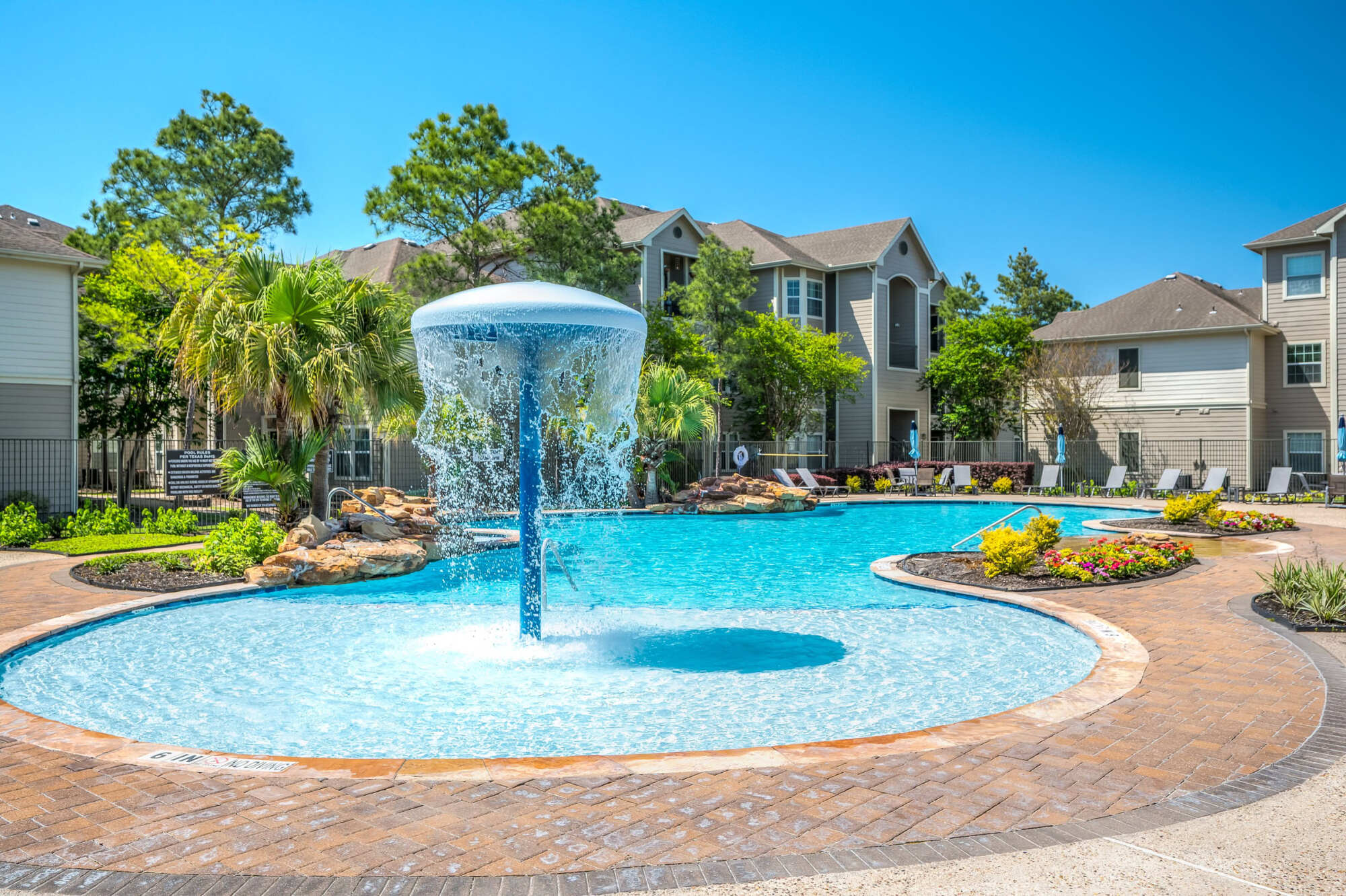 Resort-style swimming pool with a fountain feature, surrounded by lounge chairs, palm trees, and apartment buildings on a sunny day.