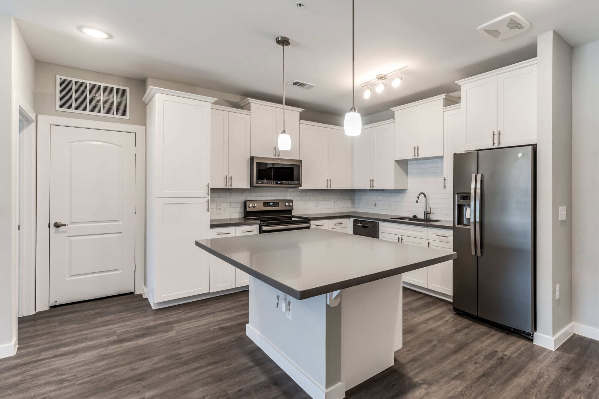 Bright apartment kitchen with white cabinetry, gray quartz island, stainless steel appliances, and pendant lights over dark wood-look flooring.