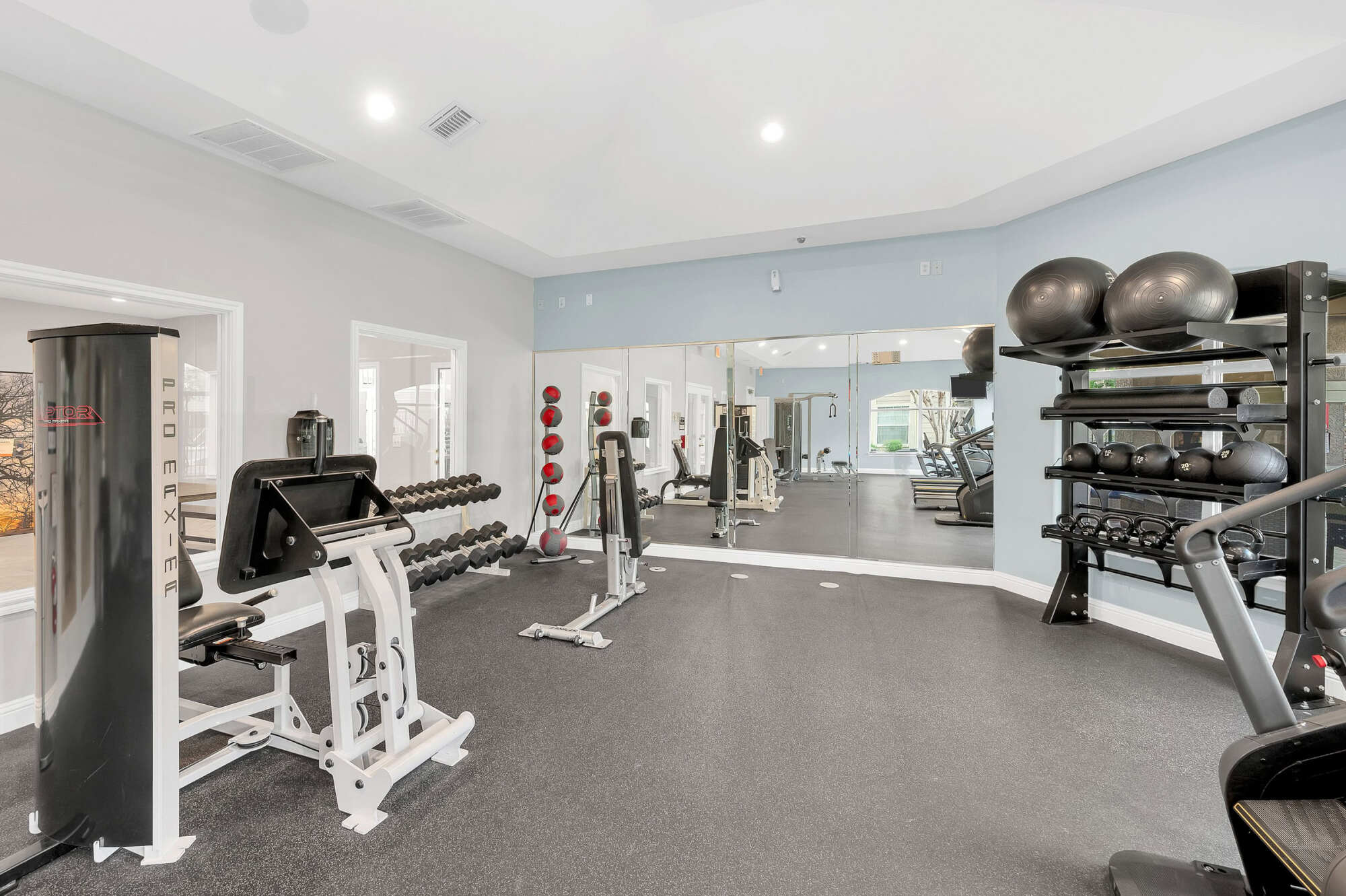 Fitness room featuring a dumbbell rack, weight machine, stability balls, mirrored wall, and cardio equipment on dark rubber flooring.