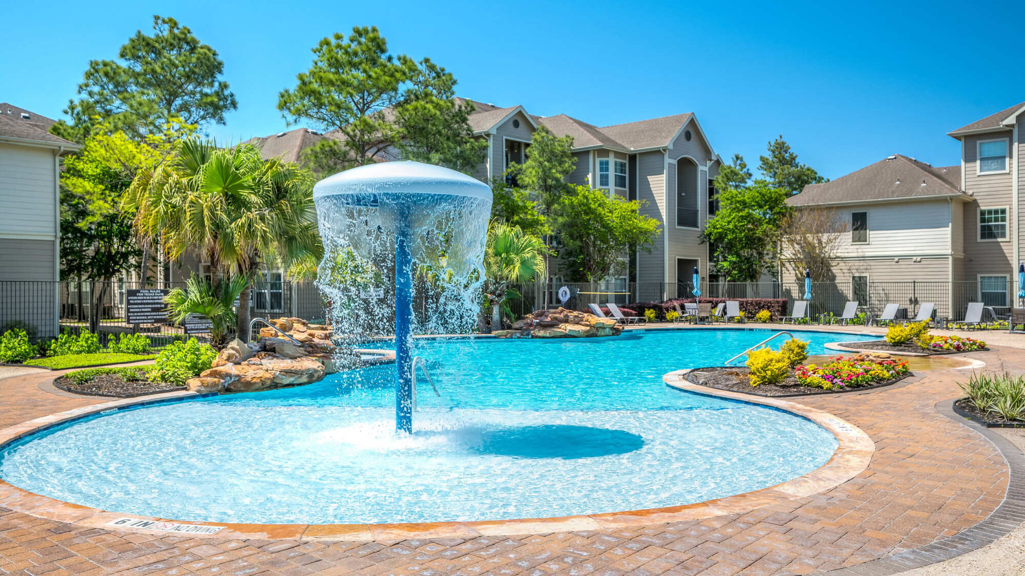 Resort-style pool with a mushroom water feature, clear blue water, lounge chairs, palm trees, and tan apartment buildings under a bright blue sky.