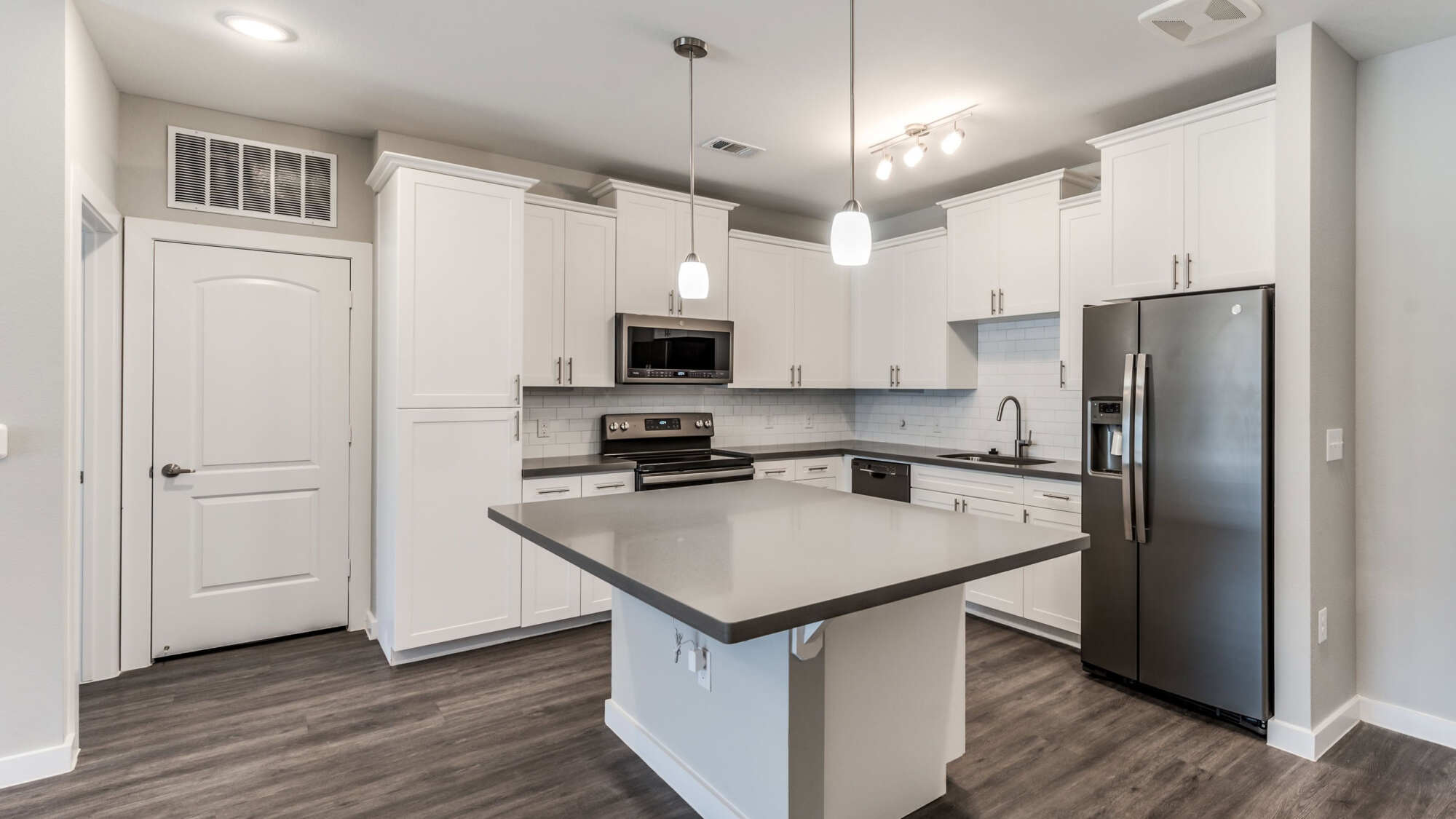 Modern apartment kitchen with white shaker cabinets, a center island with gray quartz countertop, stainless steel appliances, pendant lighting, and wood-look flooring.