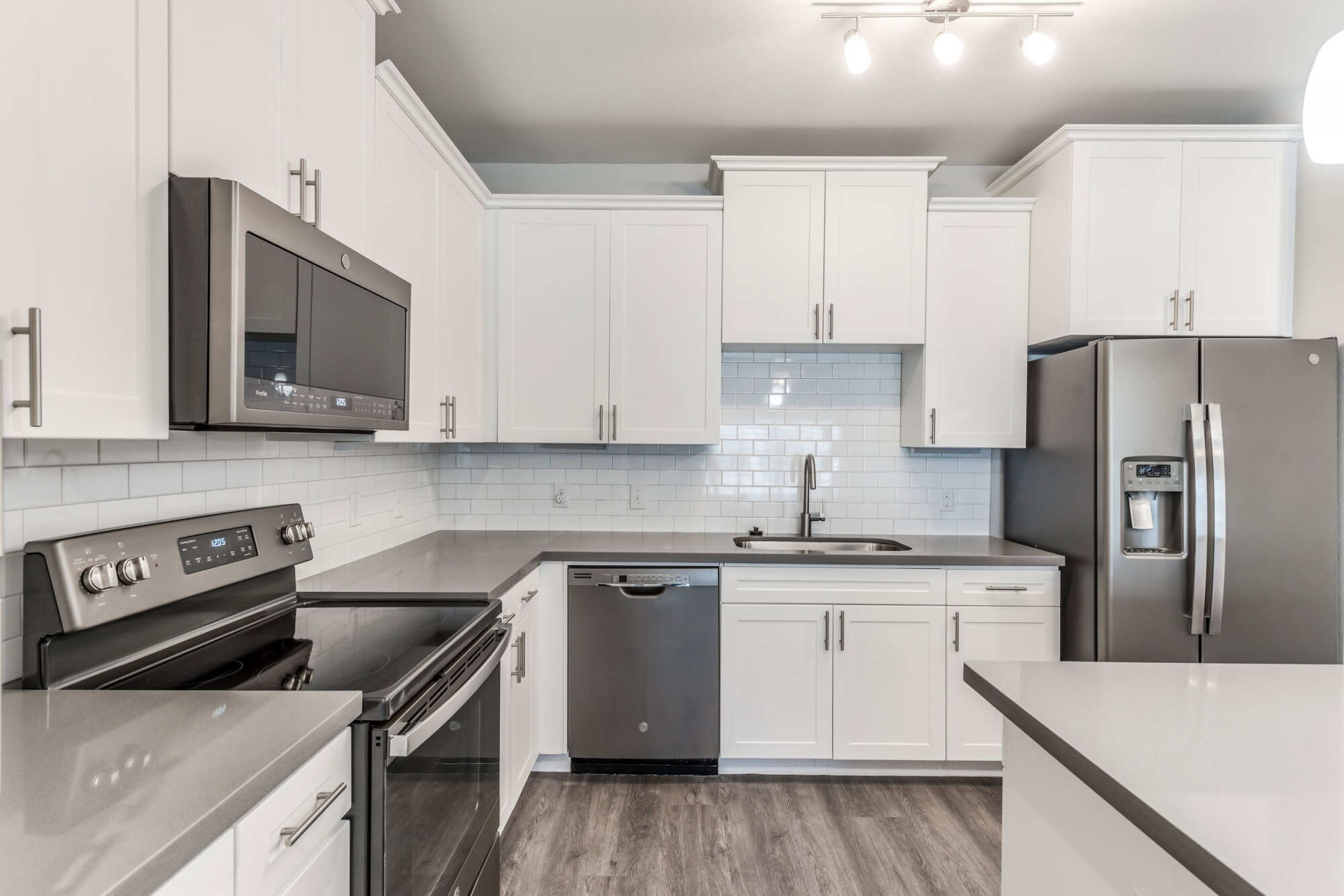 Apartment kitchen with white cabinets, gray quartz countertops, white subway tile backsplash, and stainless steel appliances including a side-by-side refrigerator.