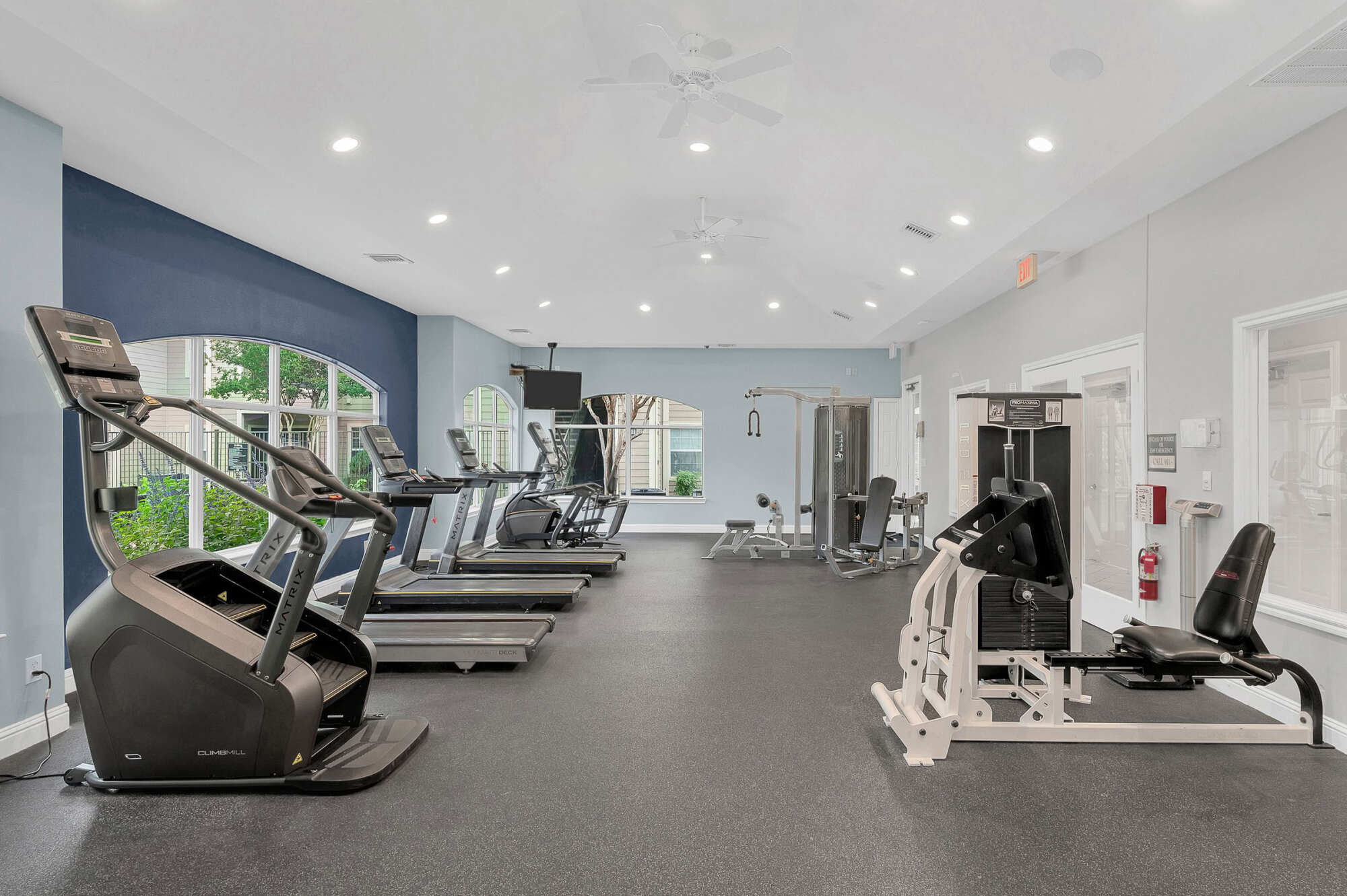 Spacious gym with a row of treadmills and a stair climber by arched windows, plus a seated weight machine, against blue and gray walls.