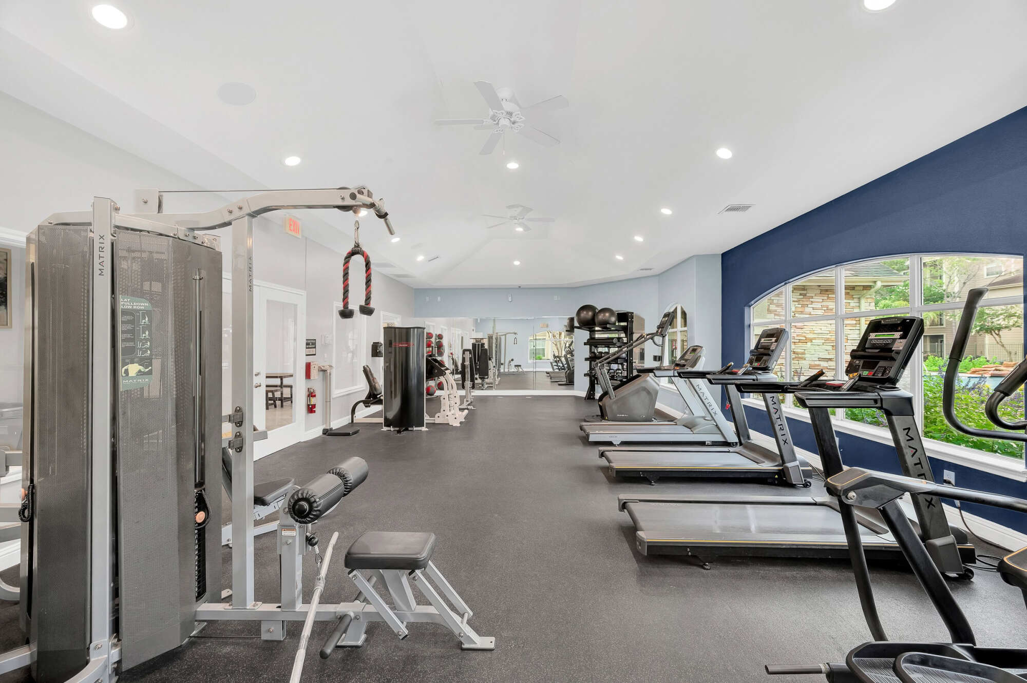 Fitness center with a cable strength machine, weight bench, treadmills, and ellipticals lined up beside large arched windows and a navy blue accent wall.