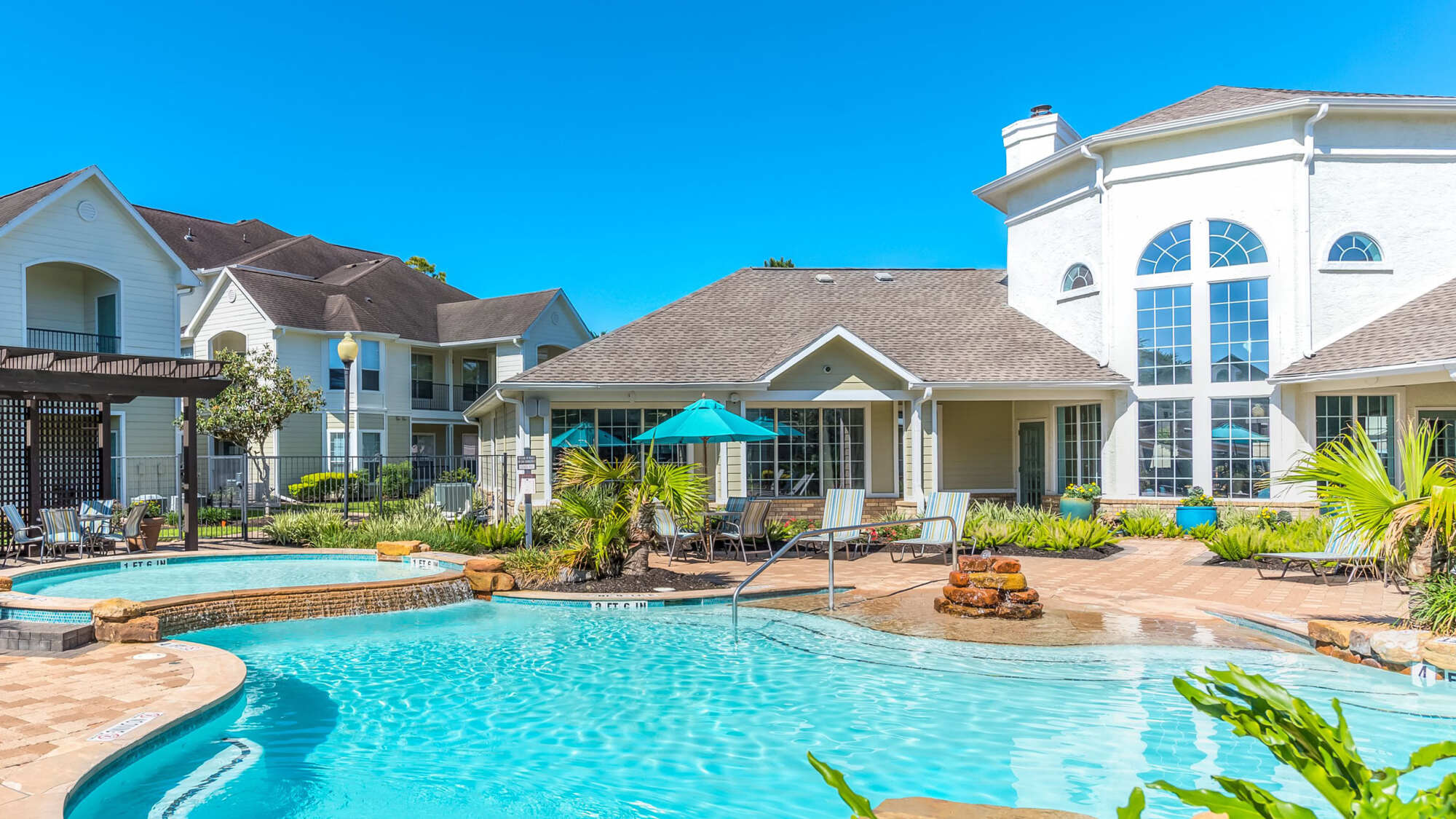 Resort-style pool with crystal-blue water, lounge chairs, and shade umbrellas surrounded by apartment buildings and greenery on a sunny day.