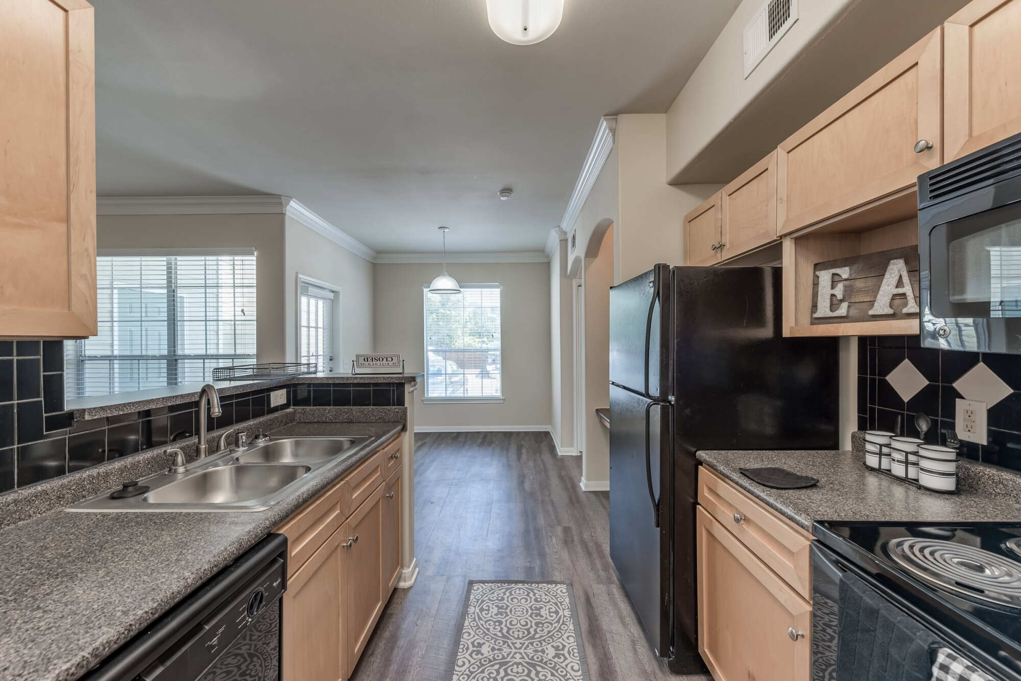 Galley-style apartment kitchen with maple cabinets, granite countertops, a stainless steel sink, black refrigerator and range, and a dining nook with a window.