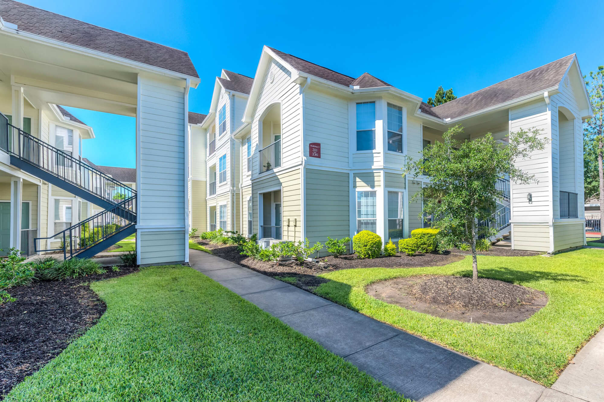 Exterior of a two-story apartment building with light siding, gabled rooflines, balconies, and an exterior staircase, set among manicured lawns and a curving walkway.