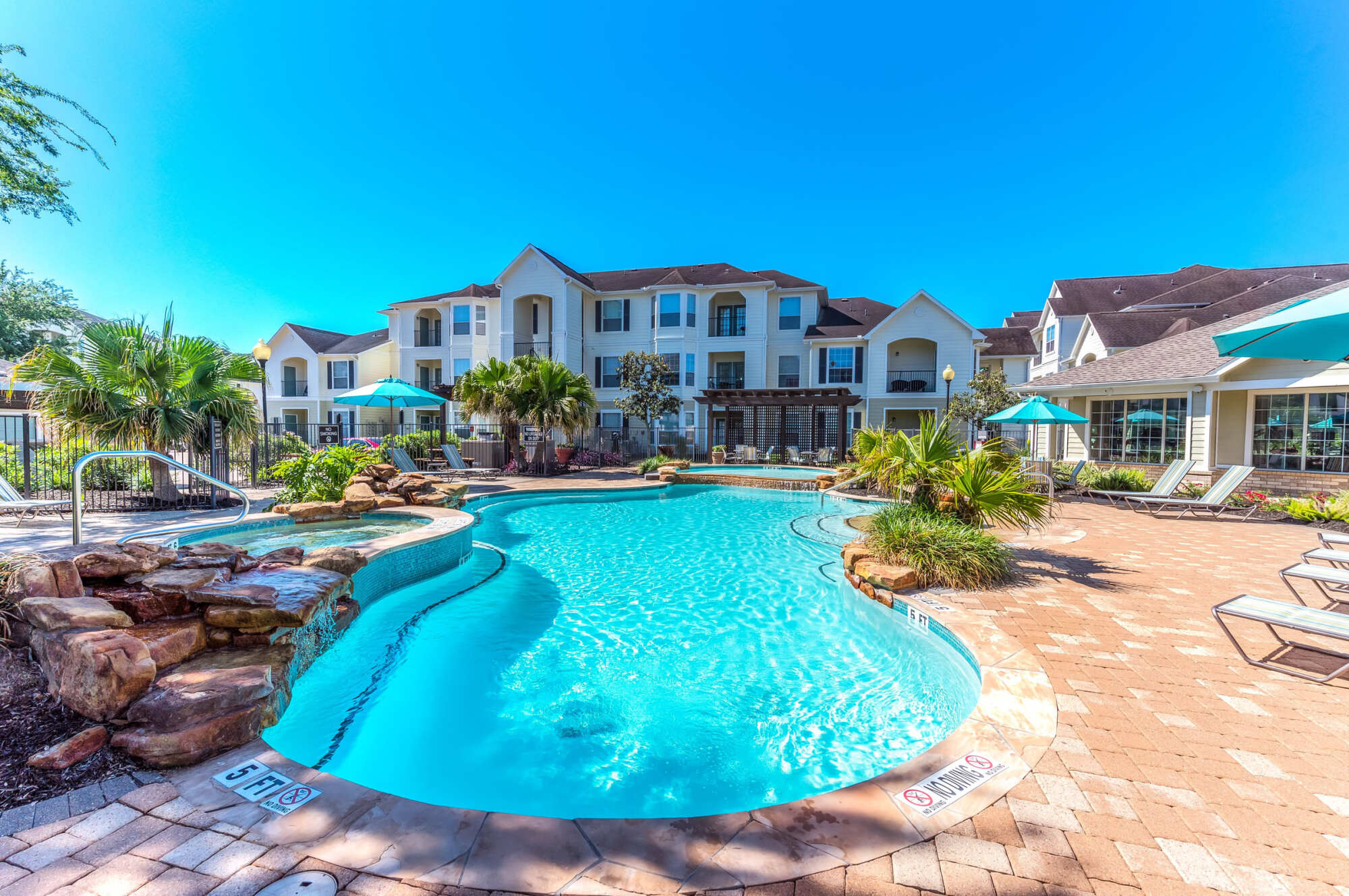Sparkling blue community pool bordered by a paved deck with lounge chairs and umbrellas, framed by apartment buildings and lush trees.