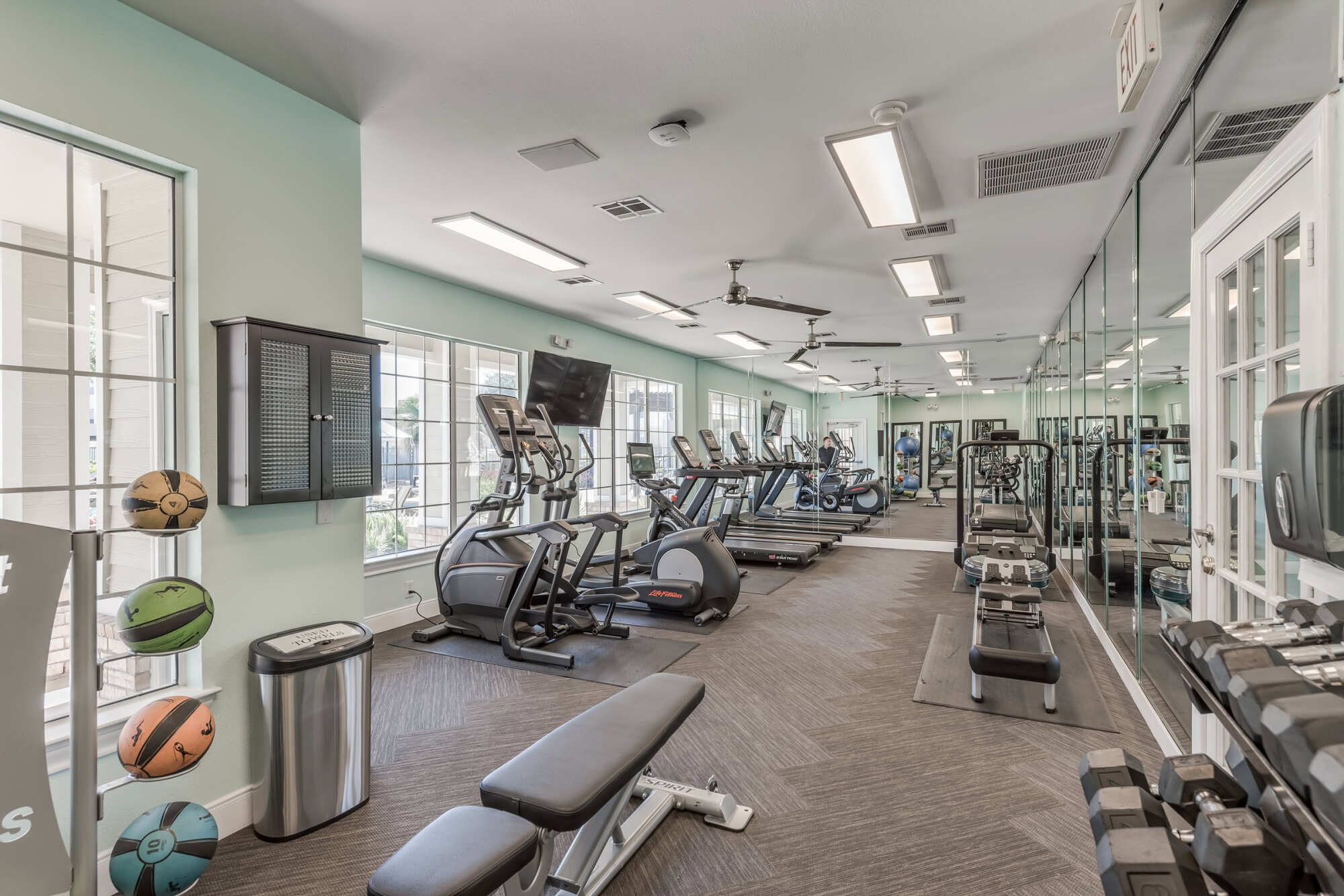 Gym area featuring weight machines, free weights, and cardio equipment along a mirrored wall under fluorescent lighting and mint-green walls.