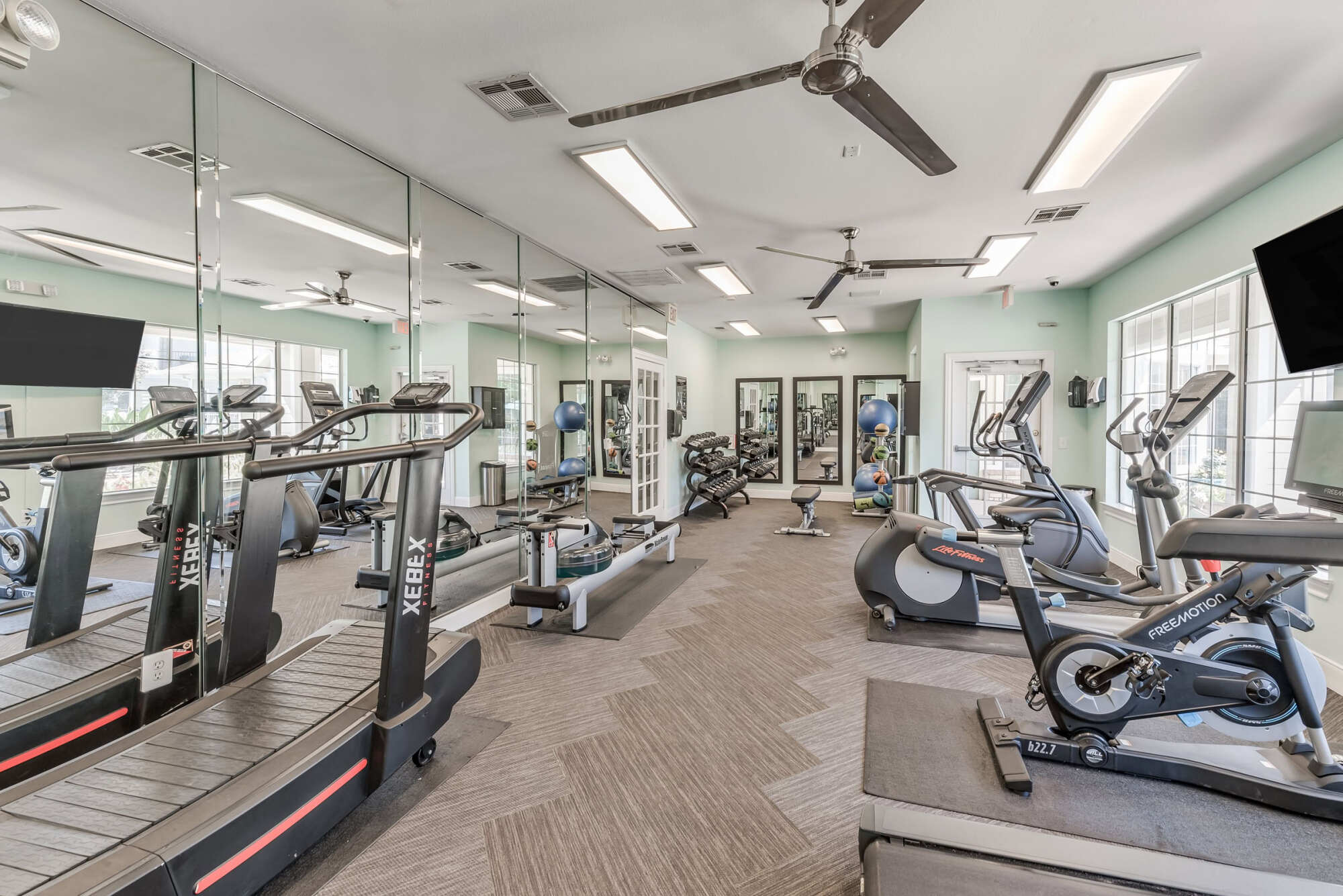 Fitness room with treadmills, a rowing machine, recumbent and spin bikes, and a mirrored wall, with windows letting in natural light.