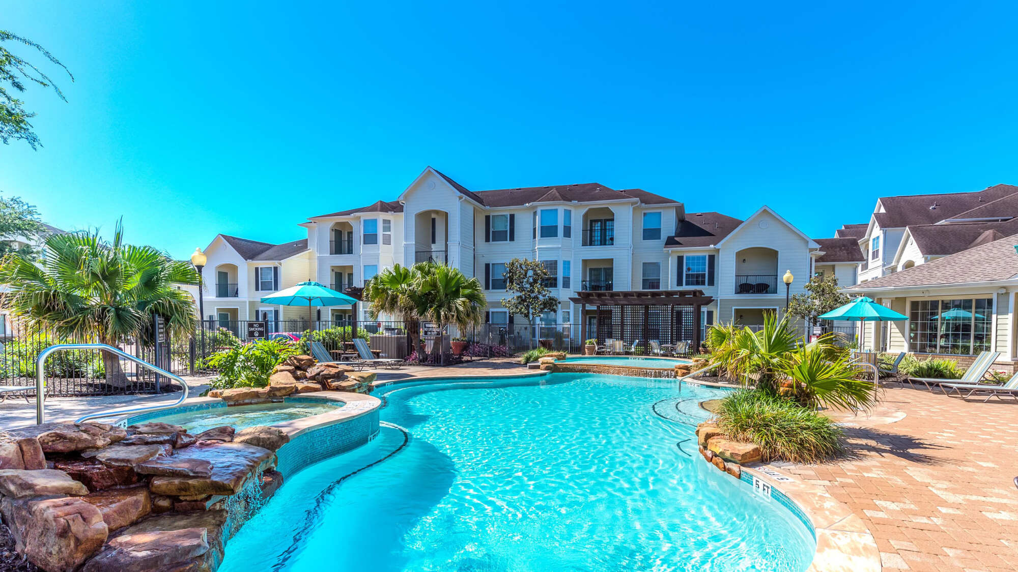Resort-style swimming pool with clear blue water, lounge chairs, shade umbrellas, and apartment buildings surrounded by greenery under a blue sky.