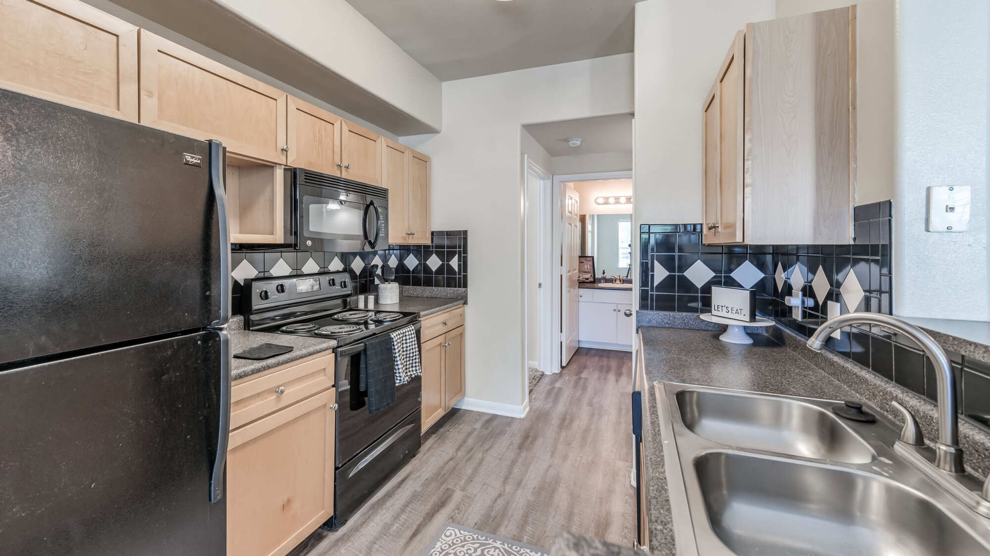 Apartment kitchen with light maple cabinets, dark granite countertops, black appliances, a tile backsplash, and a breakfast bar opening to a dining area.