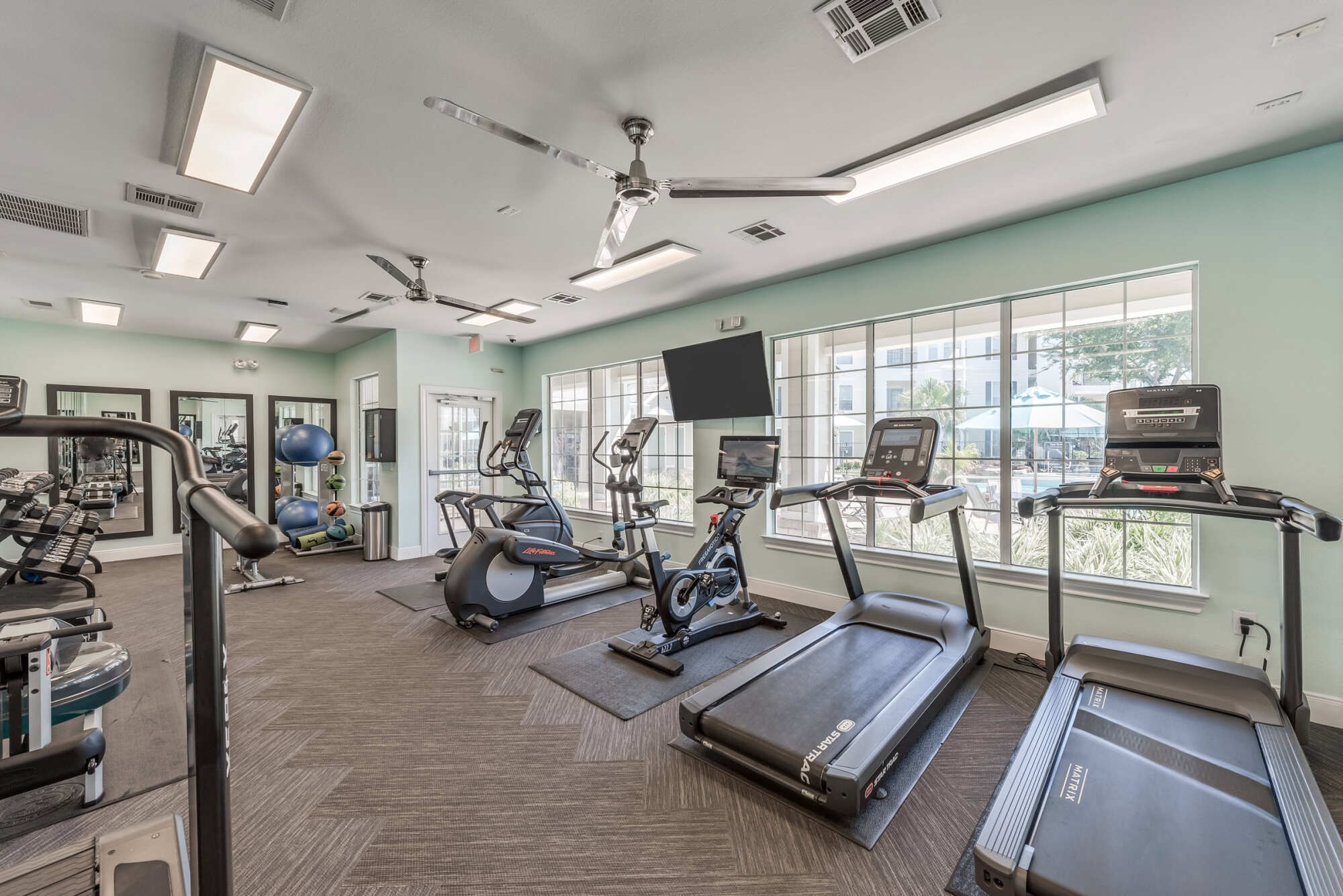 Fitness center with treadmills, an elliptical, a recumbent bike, and a mounted TV beside large windows, with a mint-green accent wall and ceiling fans.