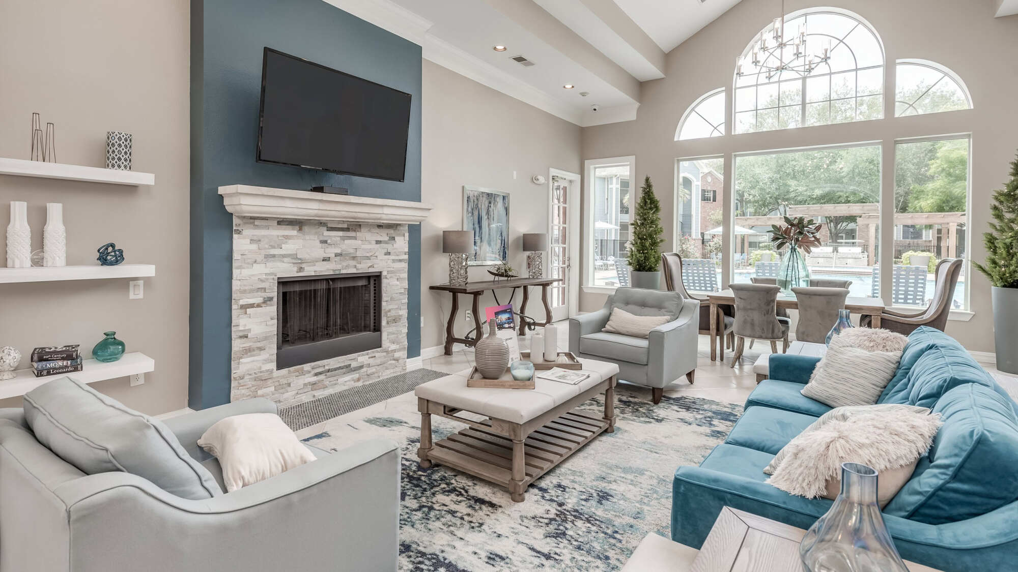 Stylishly staged clubhouse living room with a stone fireplace, mounted TV, blue and gray sofas, a dining set, and tall arched windows letting in natural light.