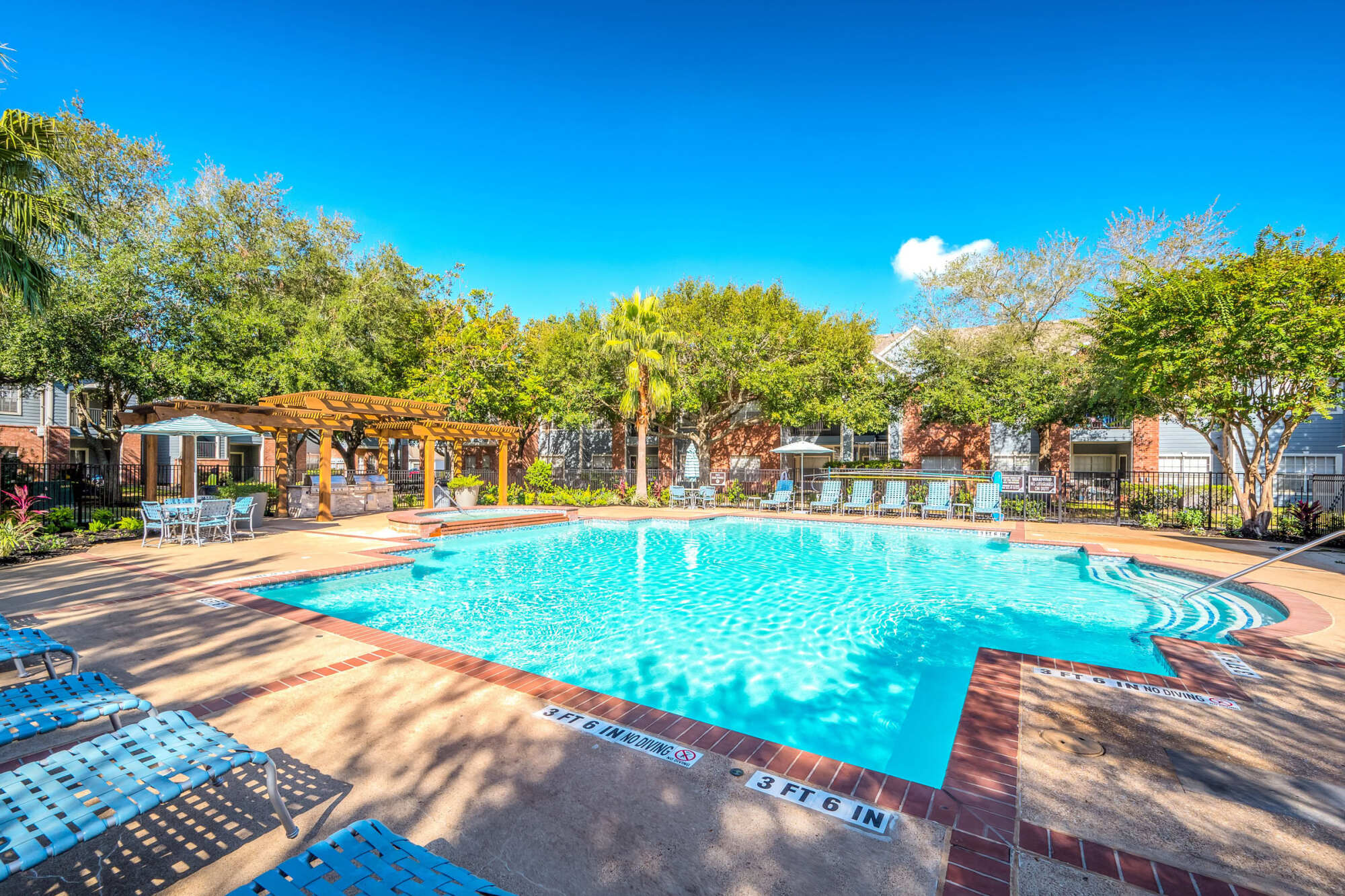 Sparkling blue community pool with a brick-trimmed deck, pergola, blue lounge chairs, and lush trees and palms surrounding the patio under a bright sky.