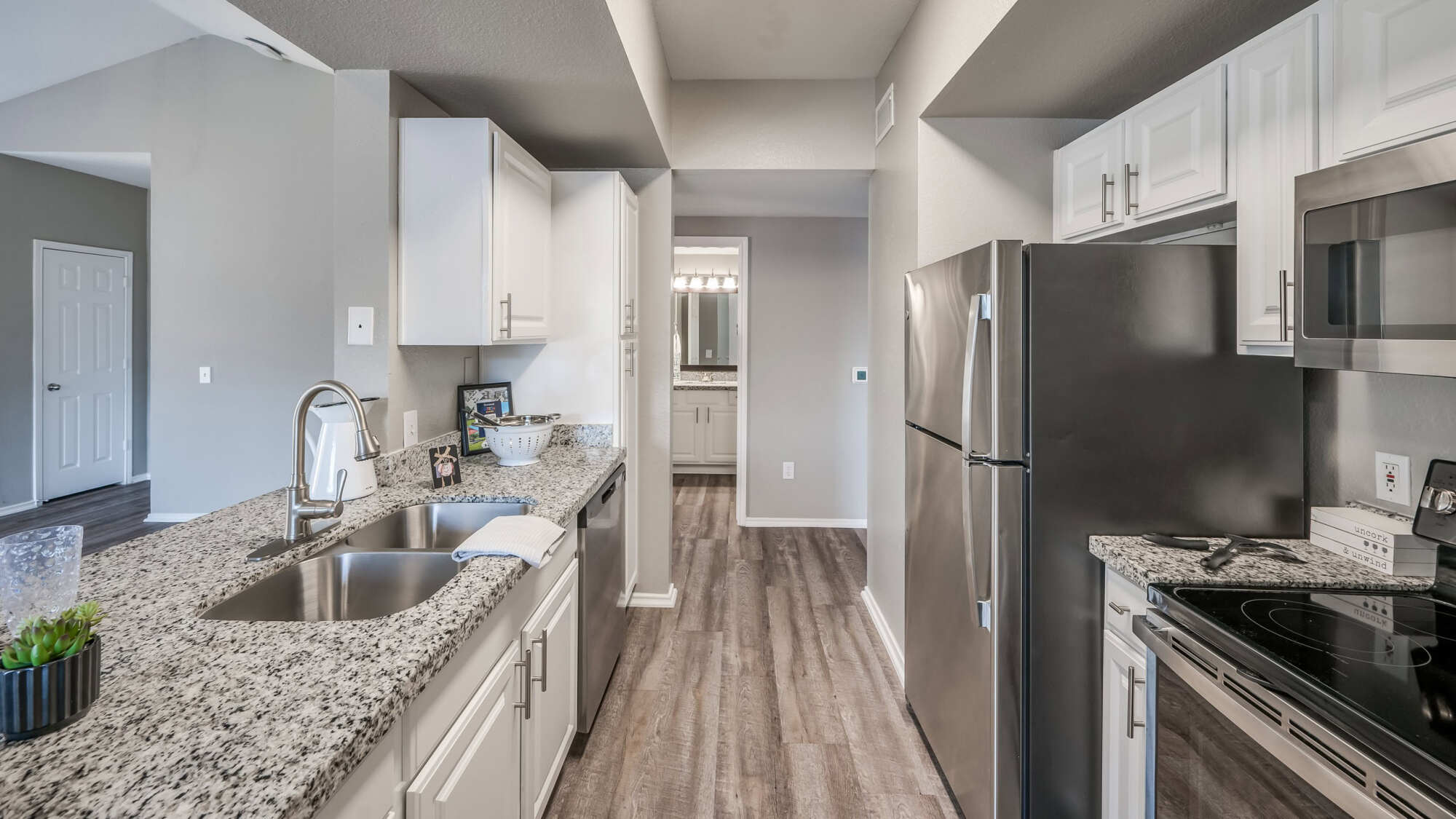 Apartment kitchen with white cabinets, speckled granite countertops, stainless steel appliances, and a breakfast bar, with a view down a hallway to the bathroom.