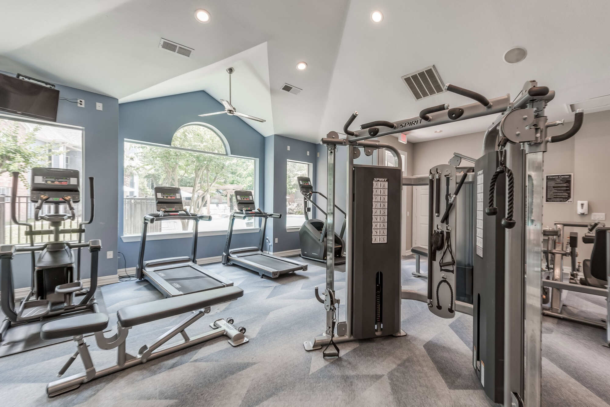 Fitness center with vaulted ceilings, arched windows, a cable strength machine, treadmills, an elliptical, and a weight bench against a blue accent wall.