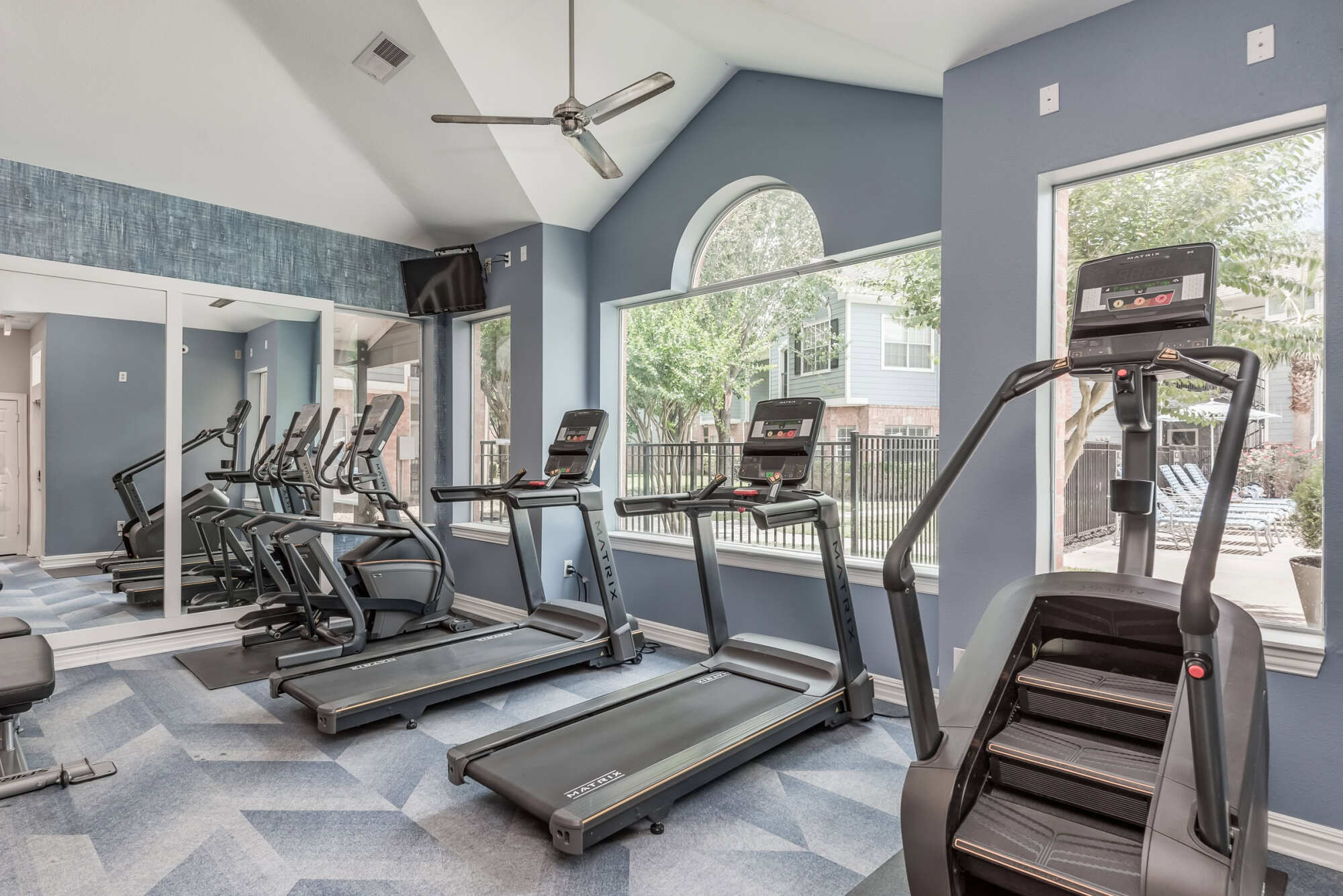Gym with a row of treadmills and a stair climber lined up beneath large arched windows, plus a mirrored wall and patterned carpet under a vaulted ceiling.