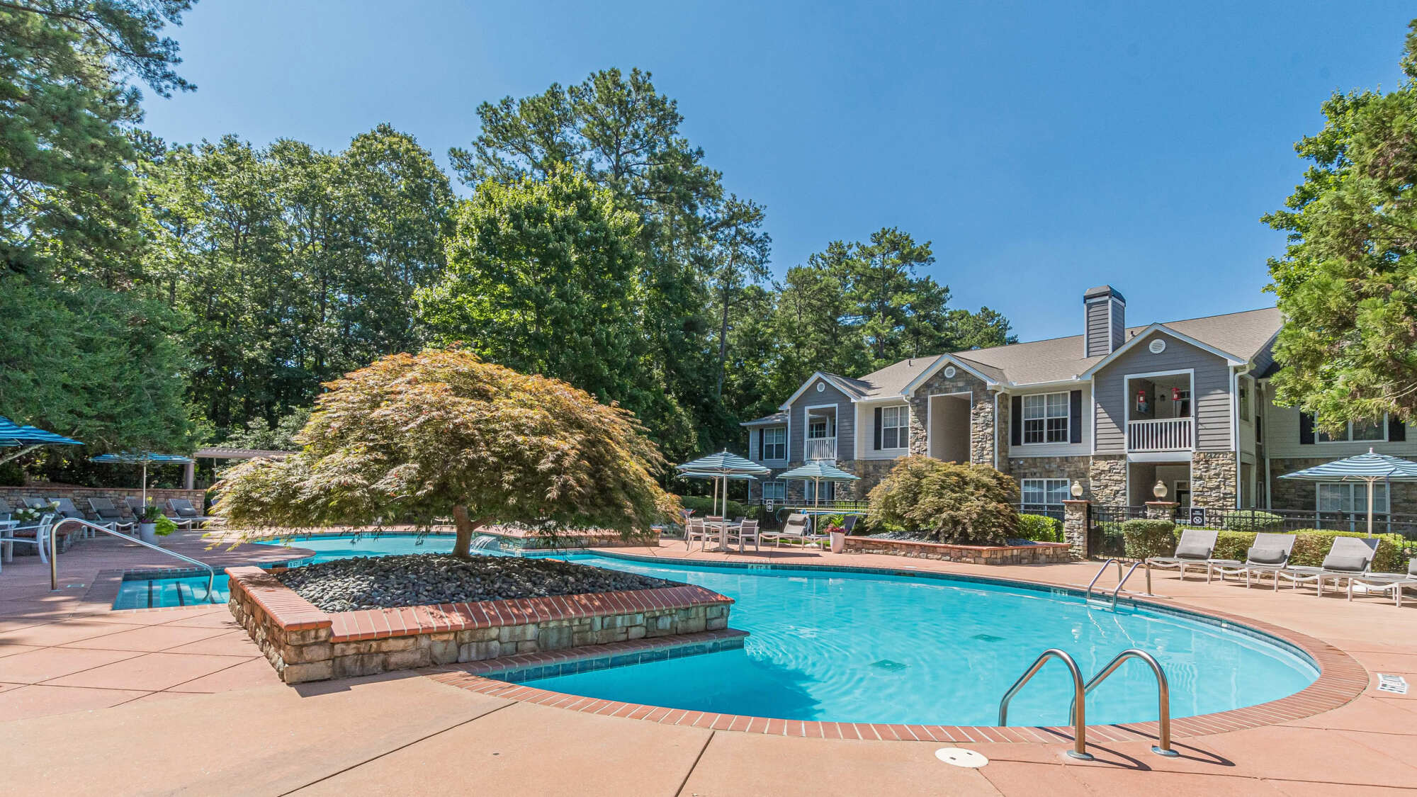 Resort-style pool with a landscaped island and Japanese maple, surrounded by a paved deck, lounge chairs, and stone-accented apartment buildings amid lush trees.