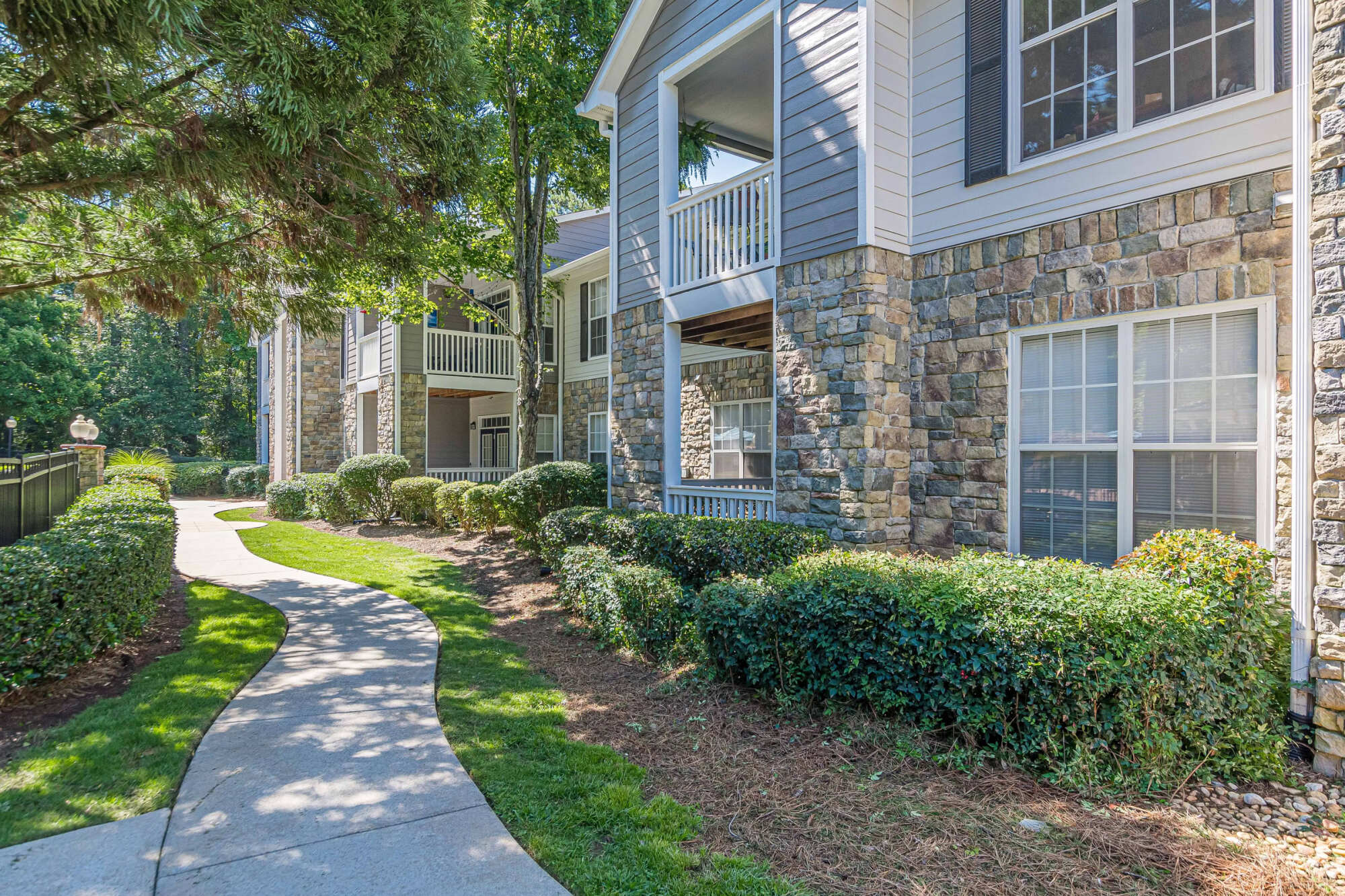 Exterior of a stone-and-siding apartment building with covered balconies, framed by manicured shrubs and a curving walkway through a shaded lawn.