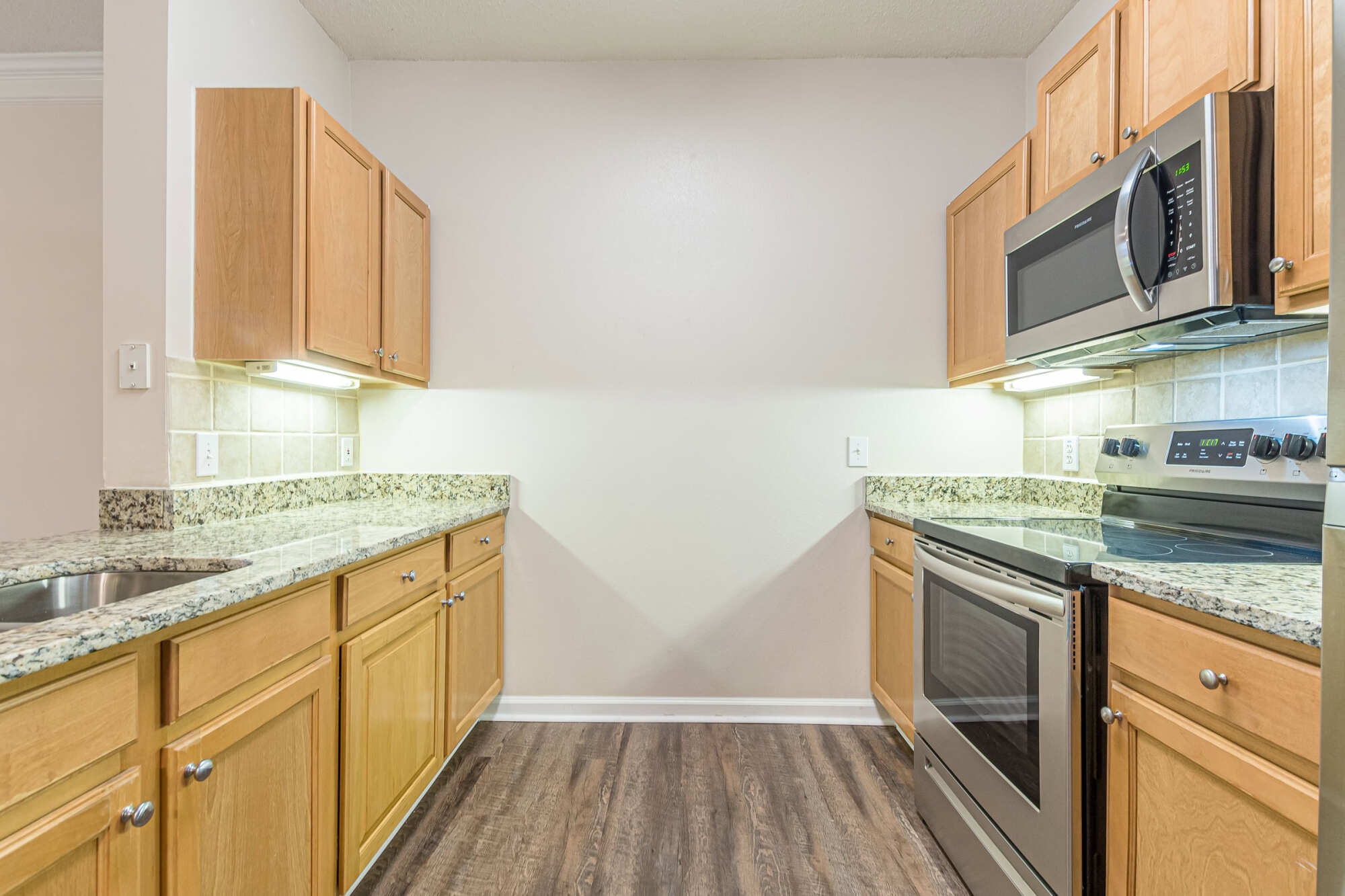 Galley kitchen with maple cabinets, granite countertops, tile backsplash, stainless steel range and over-range microwave, and wood-look flooring.