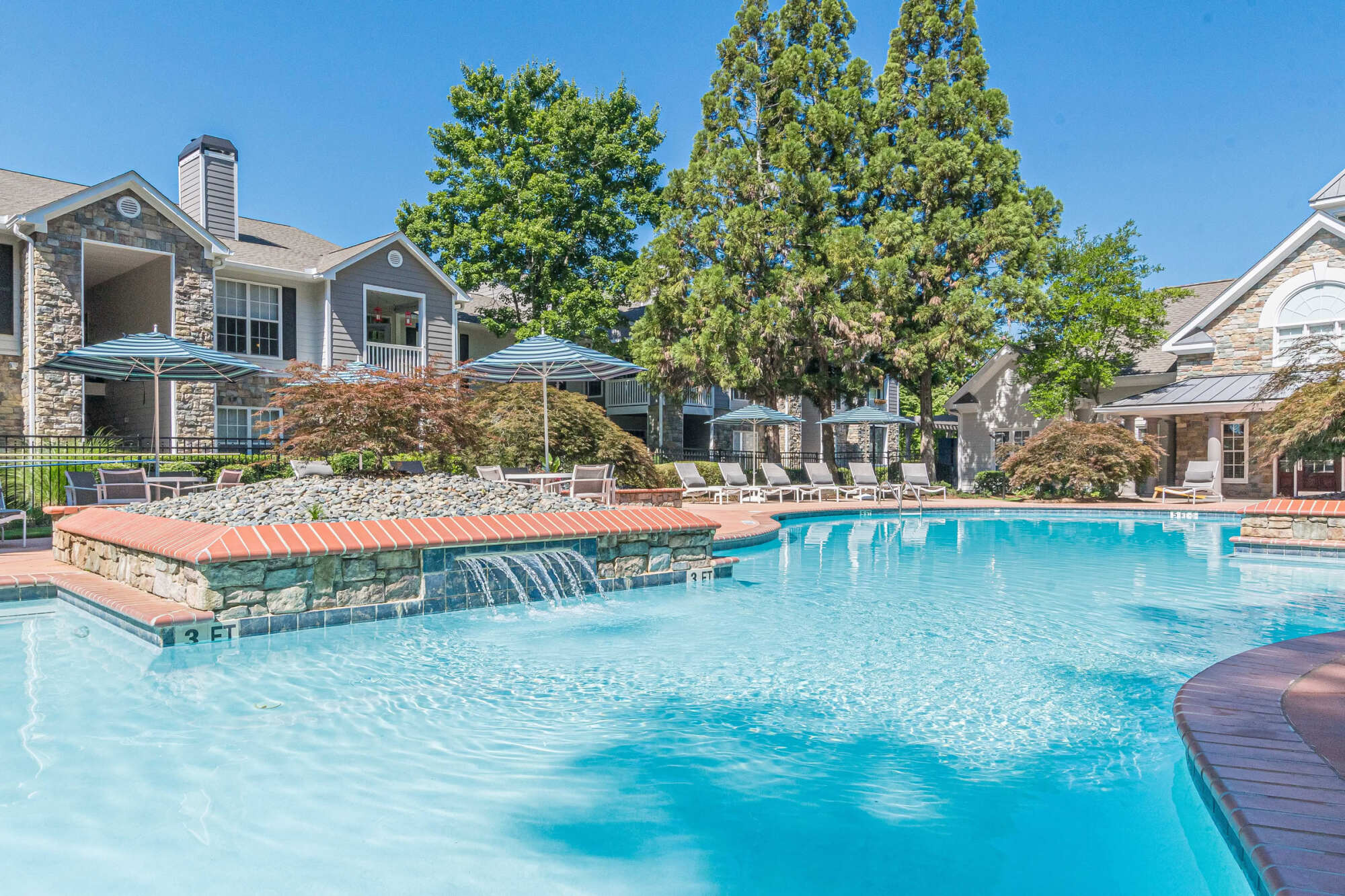 Resort-style swimming pool with a stone waterfall, brick coping, lounge chairs, and umbrellas, framed by tall trees and stone apartment buildings.