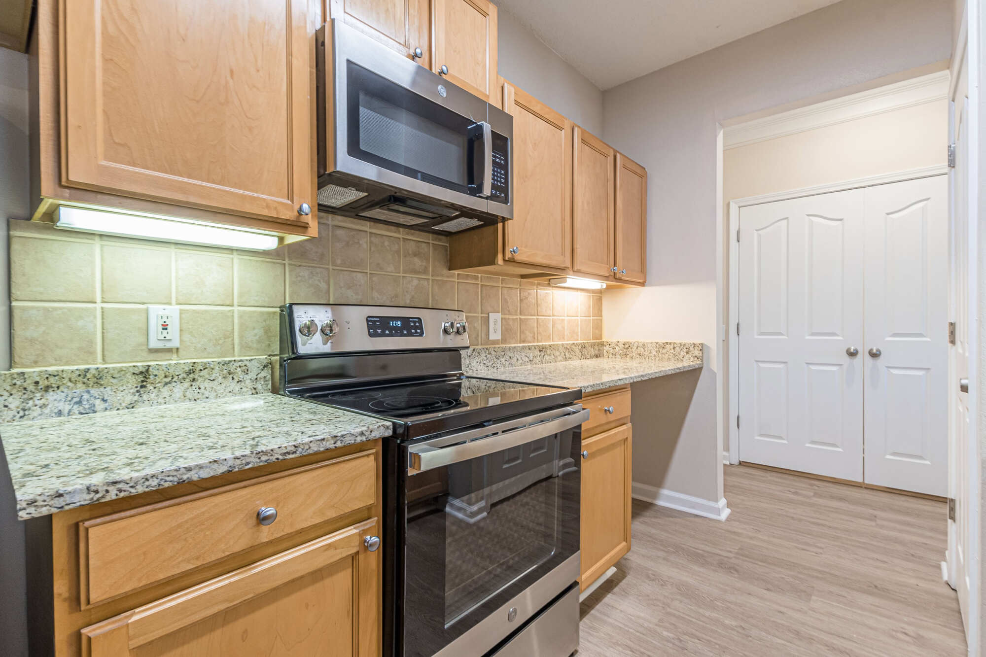 Apartment kitchen with maple cabinetry, granite countertops, tile backsplash, stainless steel range and microwave, and a white double pantry door at the end.