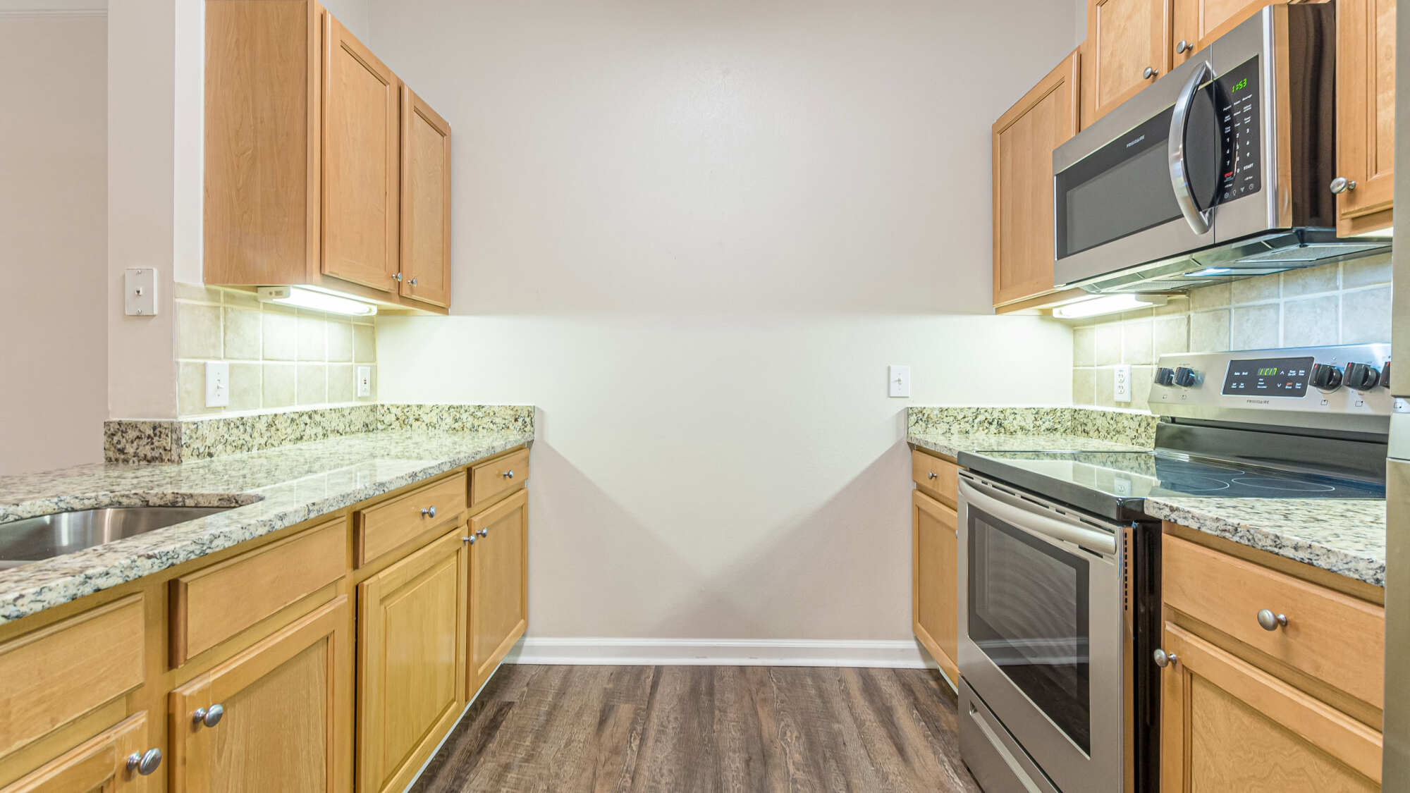 Galley kitchen with maple cabinets, speckled granite countertops, a tile backsplash, stainless steel range and over-range microwave, and wood-look flooring.