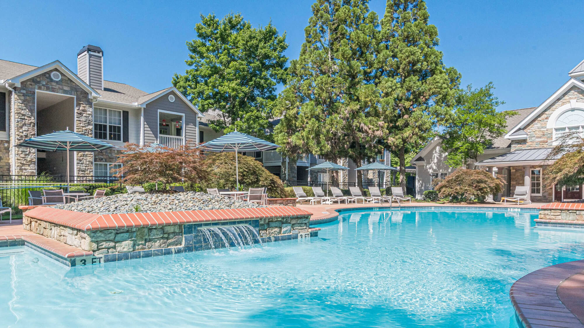 Resort-style pool with a stone waterfall feature, surrounded by lounge chairs, blue umbrellas, mature trees, and stone-and-siding apartment buildings under a blue sky.