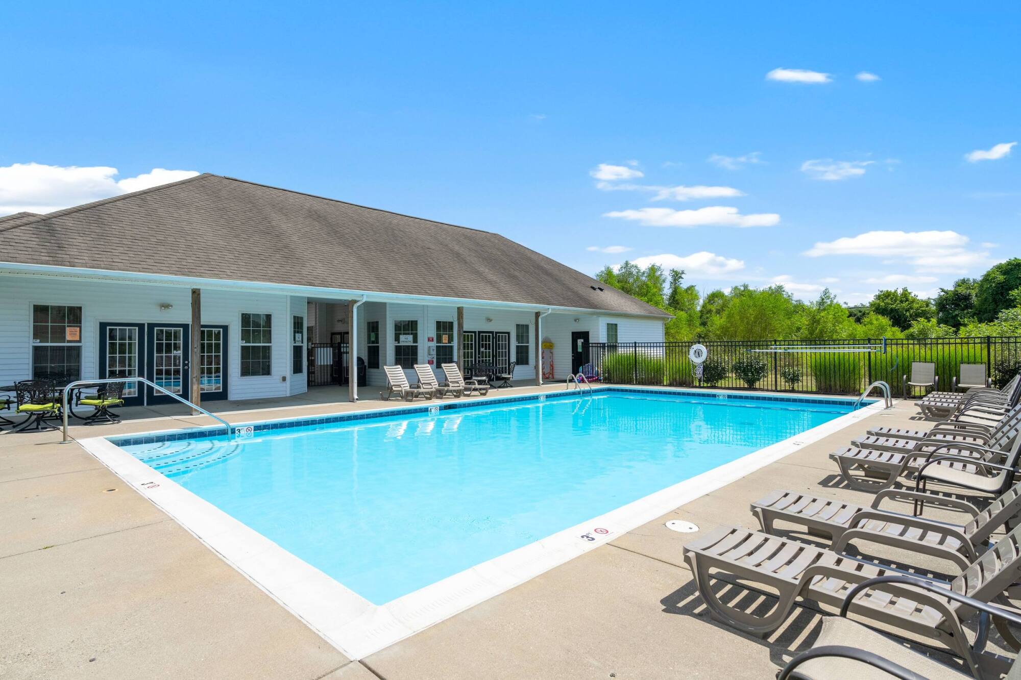 Outdoor swimming pool with lounge chairs, a fenced area, and a white clubhouse under a clear blue sky.