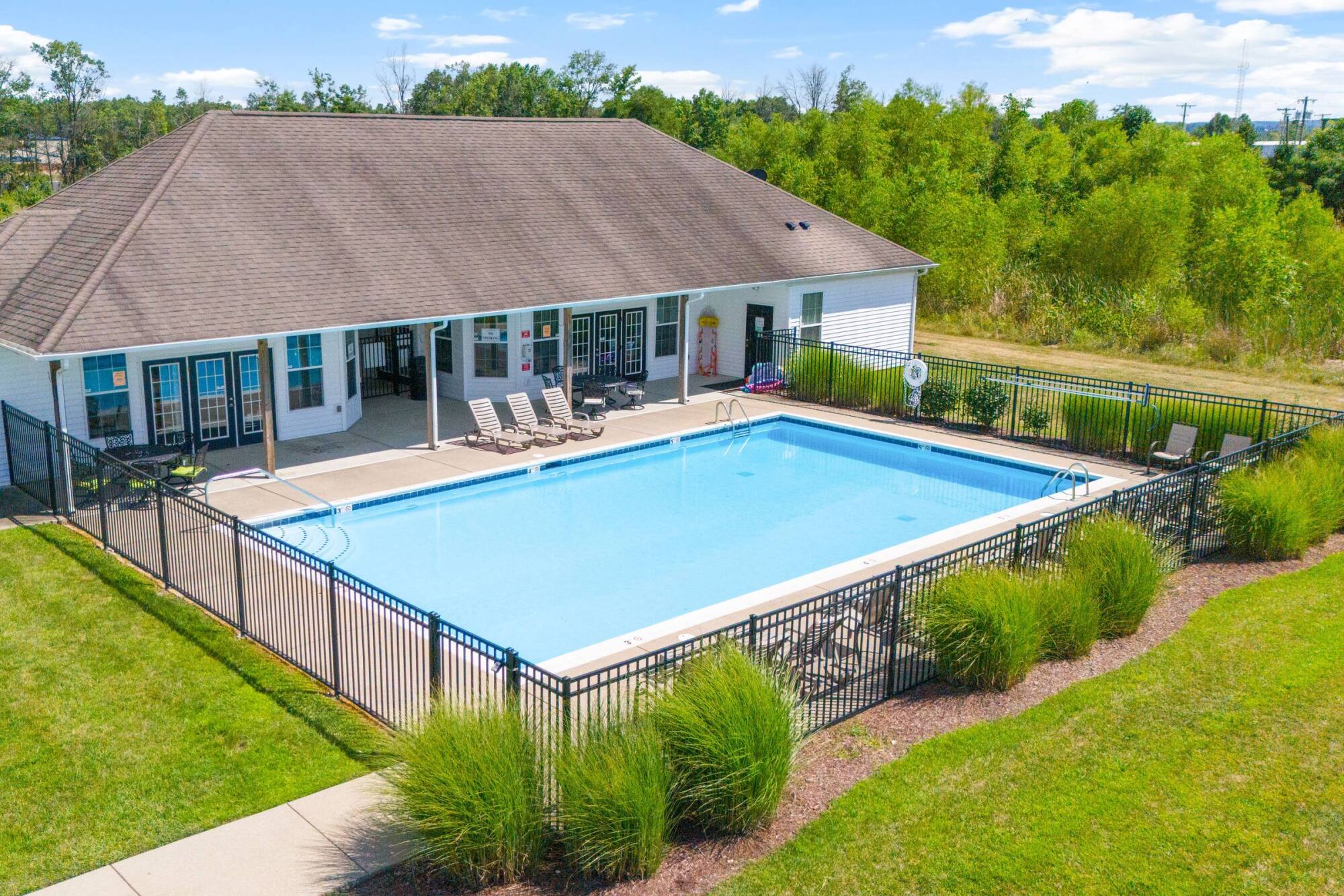 A fenced outdoor swimming pool next to a building with lounge chairs, surrounded by green grass and trees.