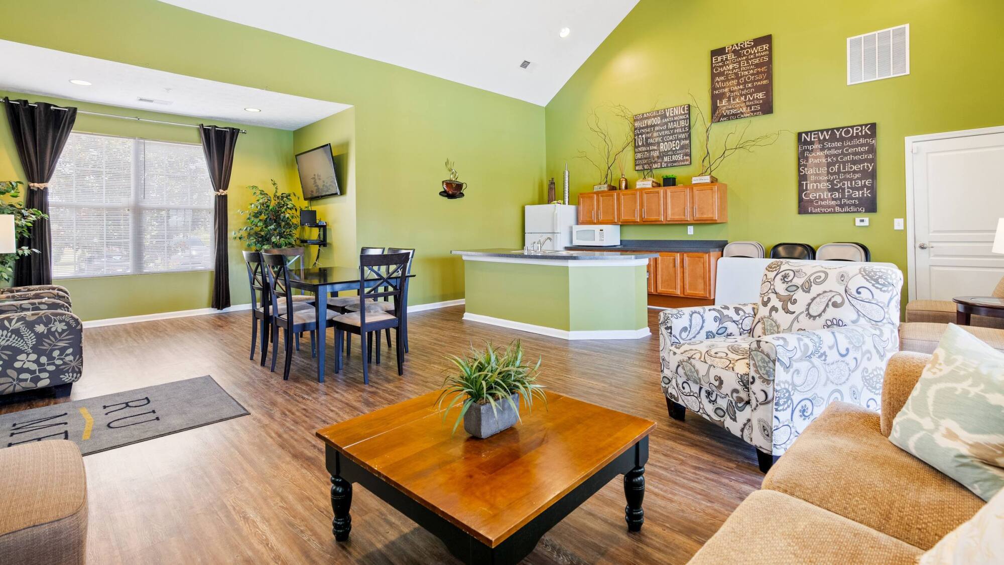 Bright living-dining area with green walls, wood floors, kitchen, wall art, and cozy seating near a large window.