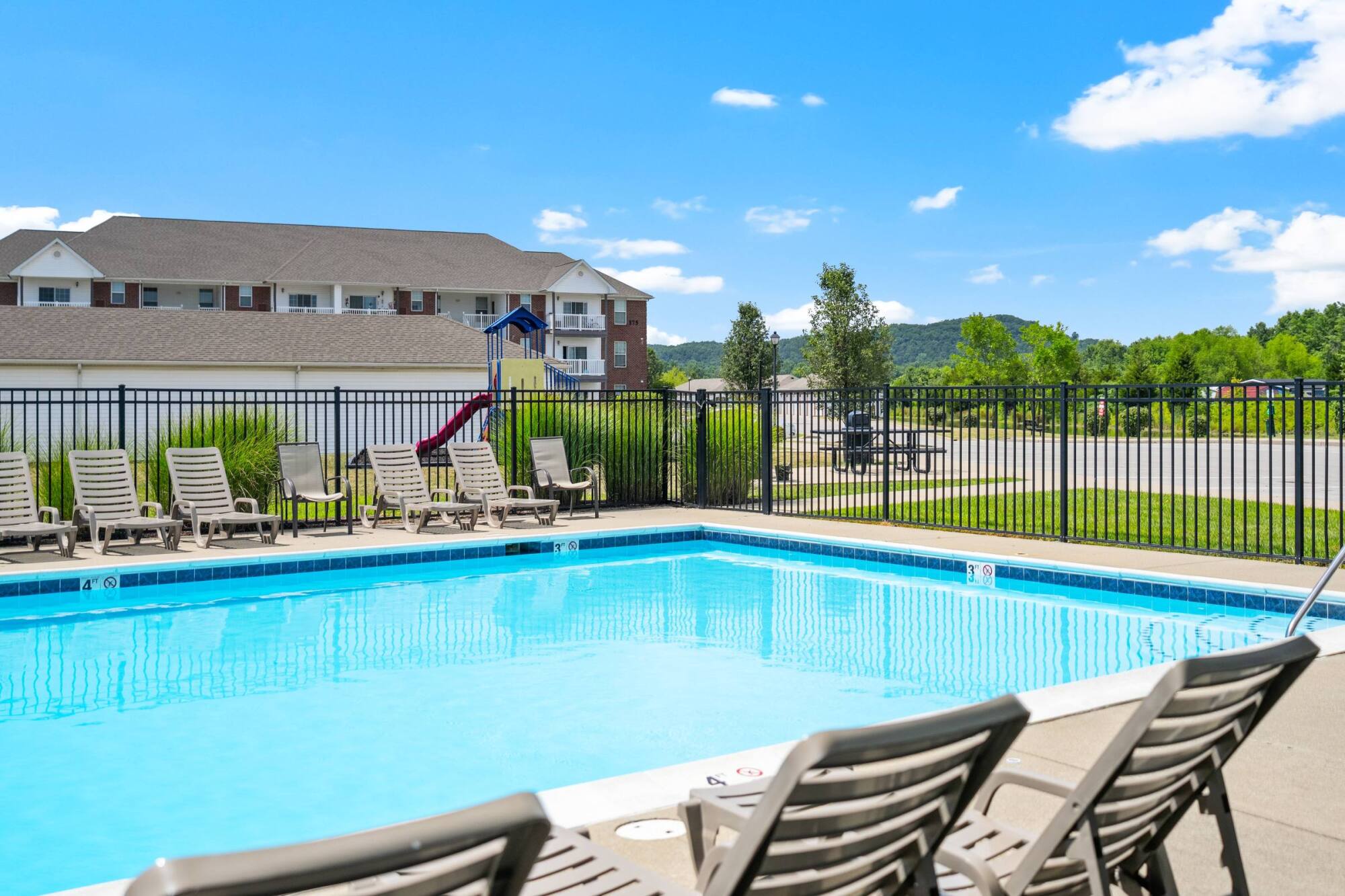 Outdoor swimming pool with lounge chairs, black metal fence, and apartment building in the background on a sunny day.