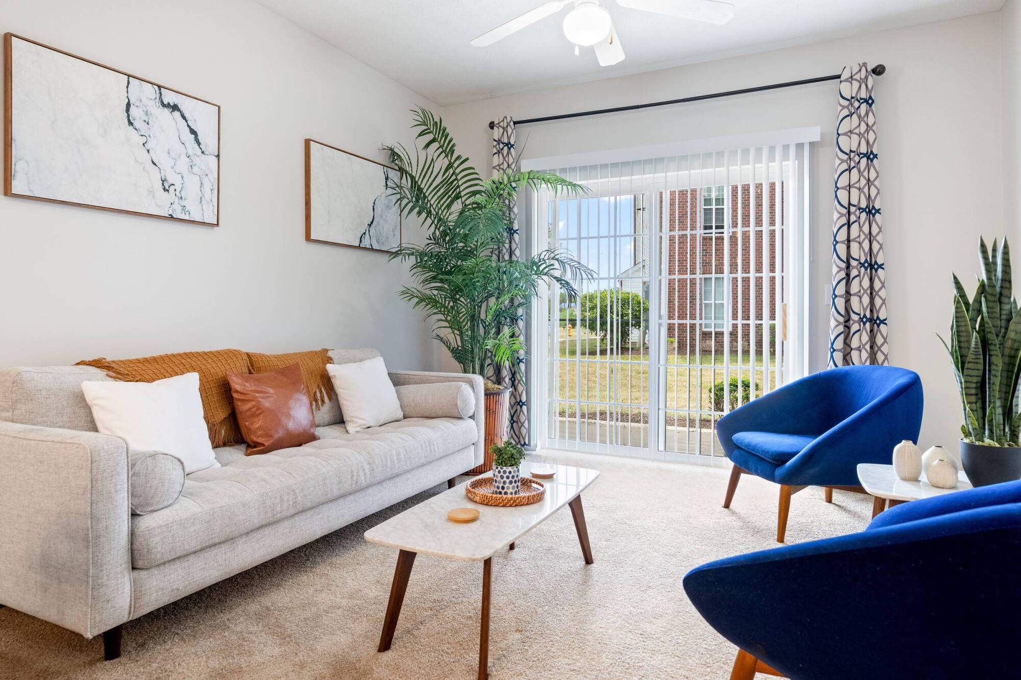 Bright living room with a beige sofa, blue chairs, plants, a marble table, and large window with vertical blinds.
