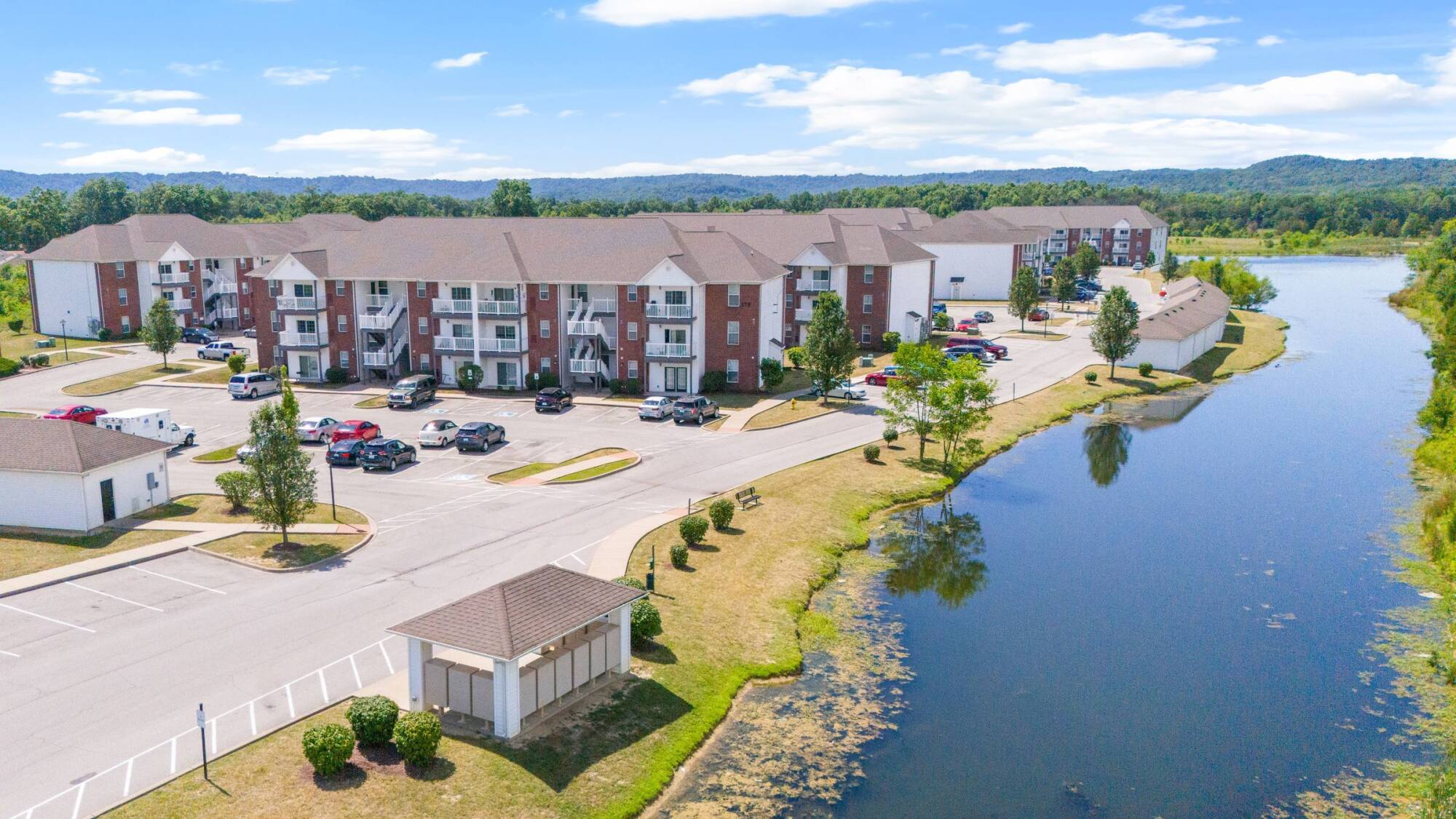 Aerial view of a lakeside apartment complex with parking lot, cars, and green trees under a blue sky.