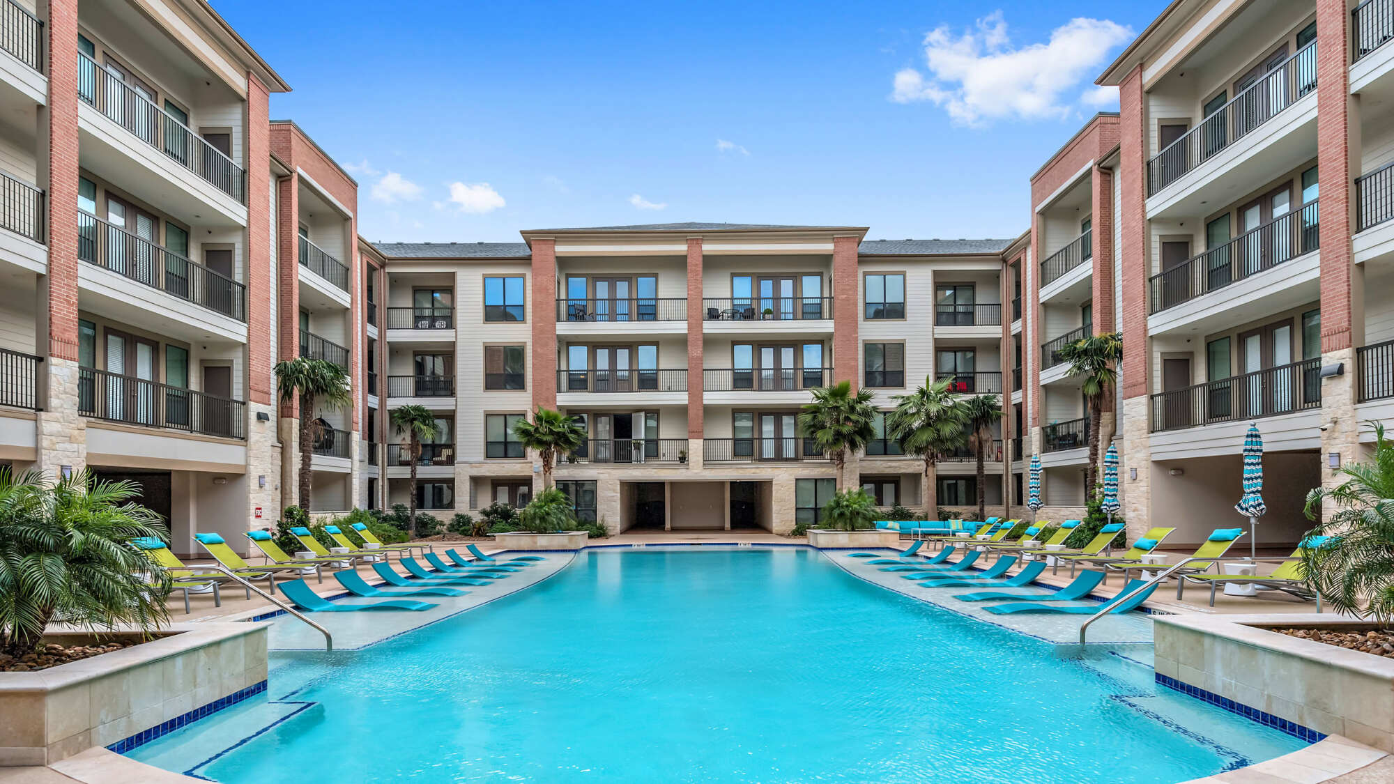 Courtyard swimming pool flanked by colorful lounge chairs and palm trees, framed by a U-shaped multi-story apartment building under a clear blue sky.