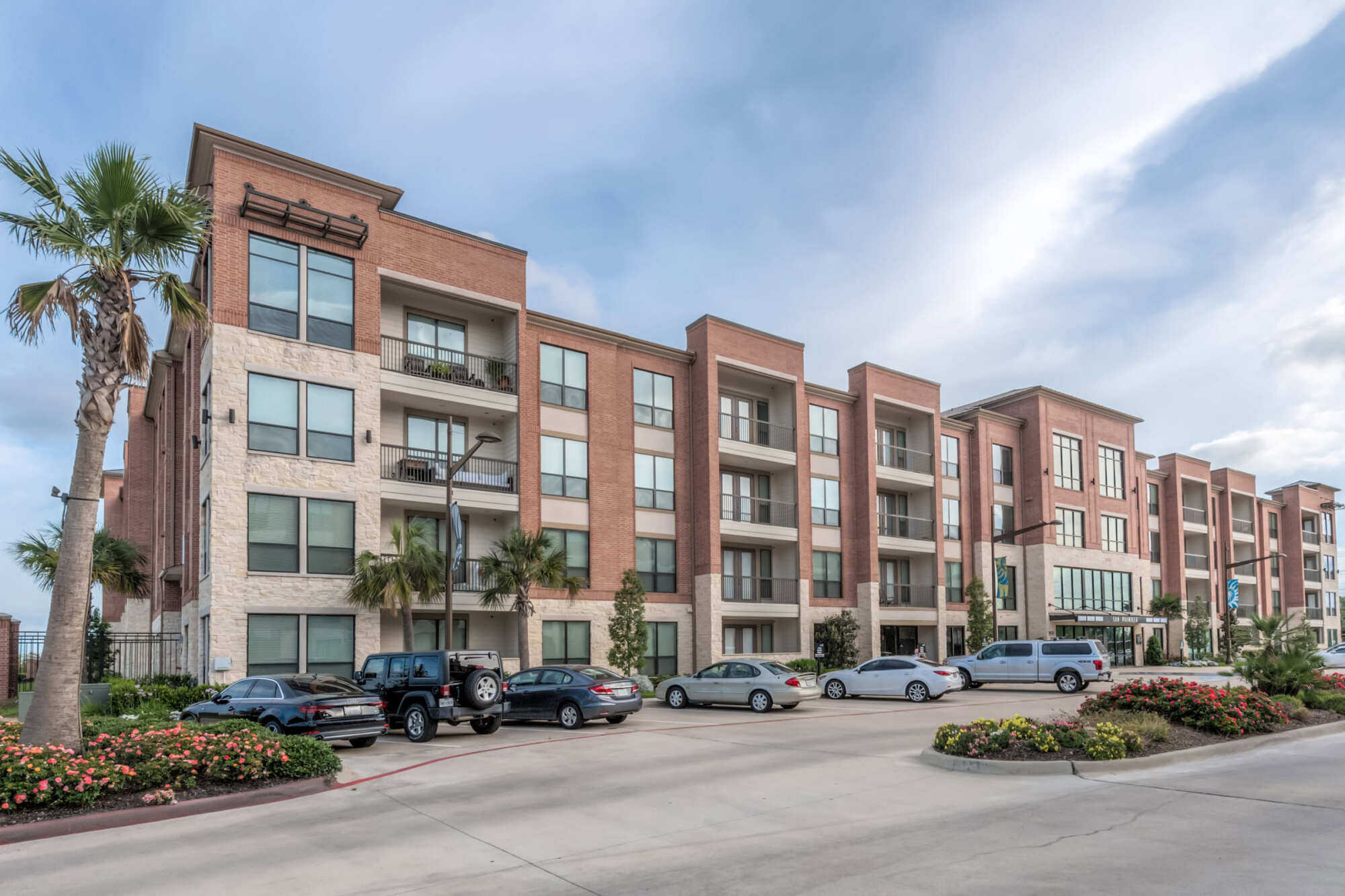 Exterior of a four-story brick-and-stucco apartment community with balconies, palm trees, and a parking area with cars under a partly cloudy sky.