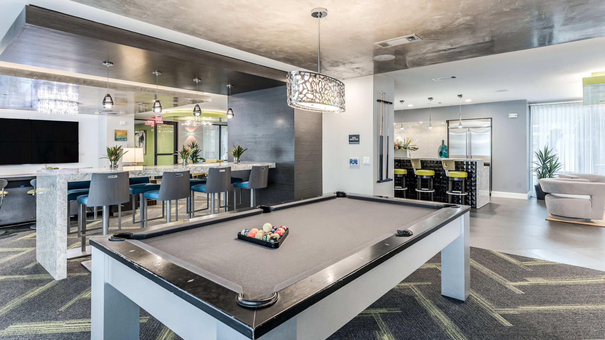 Modern clubhouse lounge with a gray-felt billiards table, a kitchen and bar with blue stools, a drum-shade chandelier, and contemporary seating areas.
